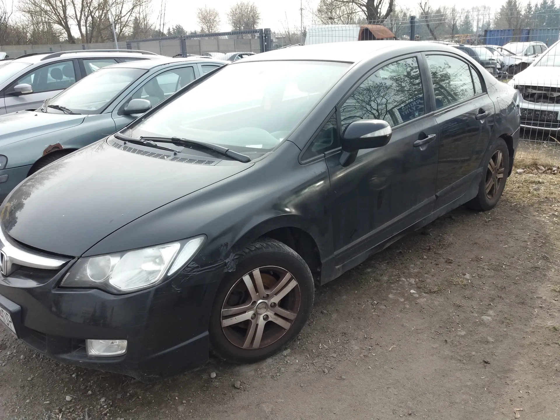 Czarny samochód osobowy typu sedan z widocznymi uszkodzeniami na przodzie, w tym pękniętym reflektorem i uszkodzonym zderzakiem. Samochód zaparkowany na brudnej, ziemistej powierzchni. W tle inne uszkodzone pojazdy, metalowe ogrodzenie oraz drzewa bez liści. Częściowo widoczna tablica rejestracyjna z literami WOP. Niebo pochmurne.