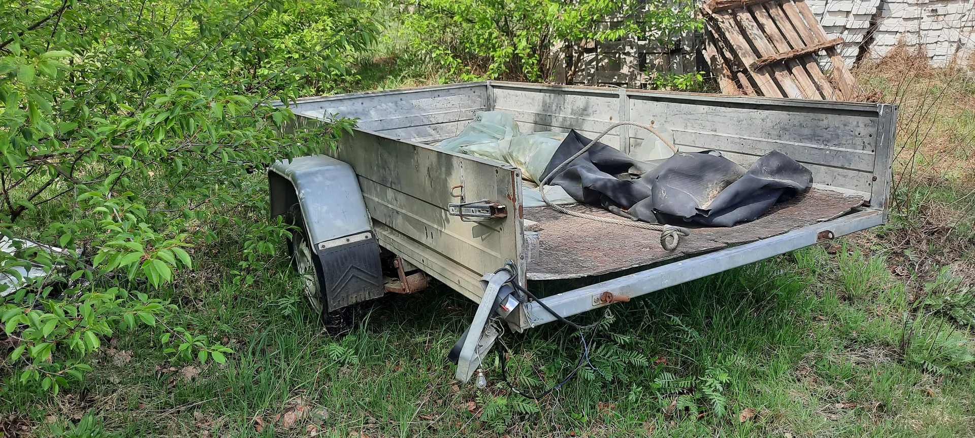 Stary metalowy przyczepa o otwartym ladowniku stojąca na trawie. W ladowniku znajdują się czarne i zielone folie oraz sznur. Przyczepa ma zniszczoną powierzchnię, widoczne są rdza i ślady zużycia. W tle rosną zielone krzewy i drzewa, a także drewniana konstrukcja i stos desek. Na przodzie przyczepy widoczne są elementy elektryczne i kable.