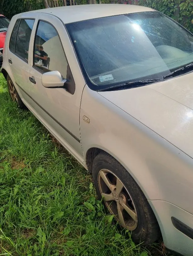 Srebrny samochód osobowy zaparkowany na trawie, widoczny z perspektywy przedniego prawego boku. Auto jest zakurzone, a w tle widać zielone krzewy i fragment innego, czerwonego pojazdu.