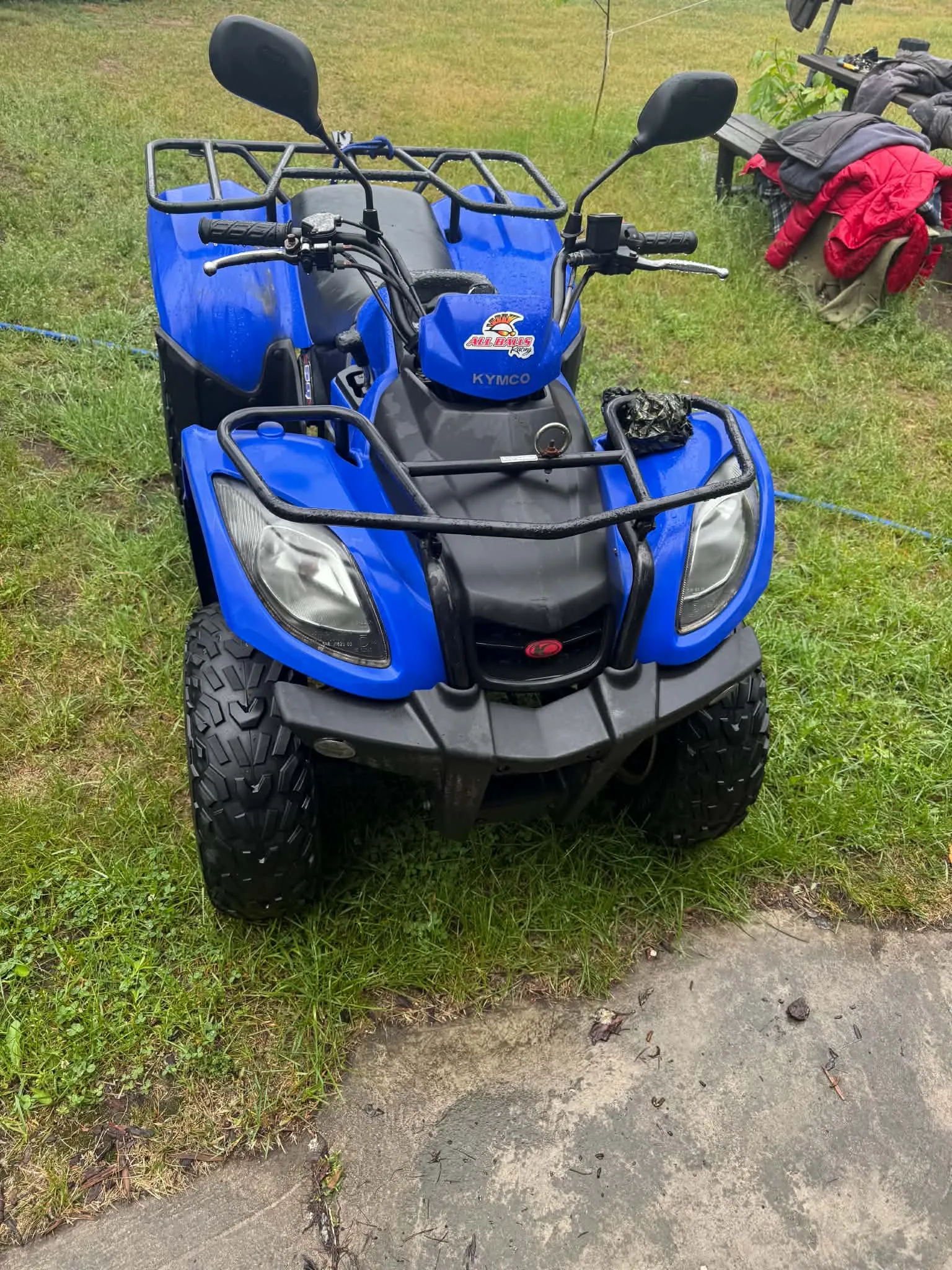 Niebieski quad marki Kymco zaparkowany na trawie. Pojazd posiada czarne opony z grubym bieżnikiem, przednią osłonę z rurek, dwa lusterka oraz bagażnik z tyłu. Na przedniej obudowie widnieje napis 'KYMCO' oraz kolorowa naklejka.