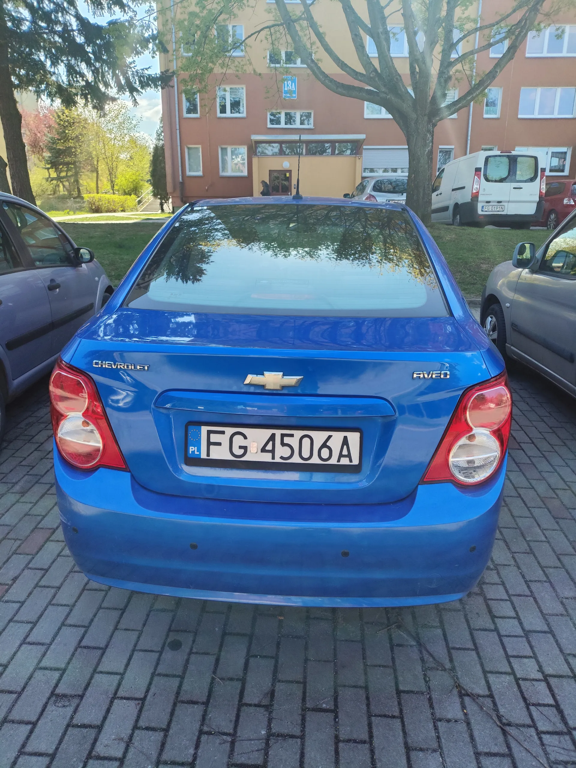 Niebieski samochód Chevrolet Aveo zaparkowany na brukowanej powierzchni. Widoczny tył pojazdu z tablicą rejestracyjną FG 4506A oraz oznaczeniem PL. Po obu stronach samochodu znajdują się inne pojazdy: szary po lewej i srebrny po prawej. W tle budynki mieszkalne o pomarańczowo-beżowej elewacji, drzewa oraz biały furgon. Na budynku widoczna tablica z numerem 13A. Niebo jest częściowo zachmurzone.