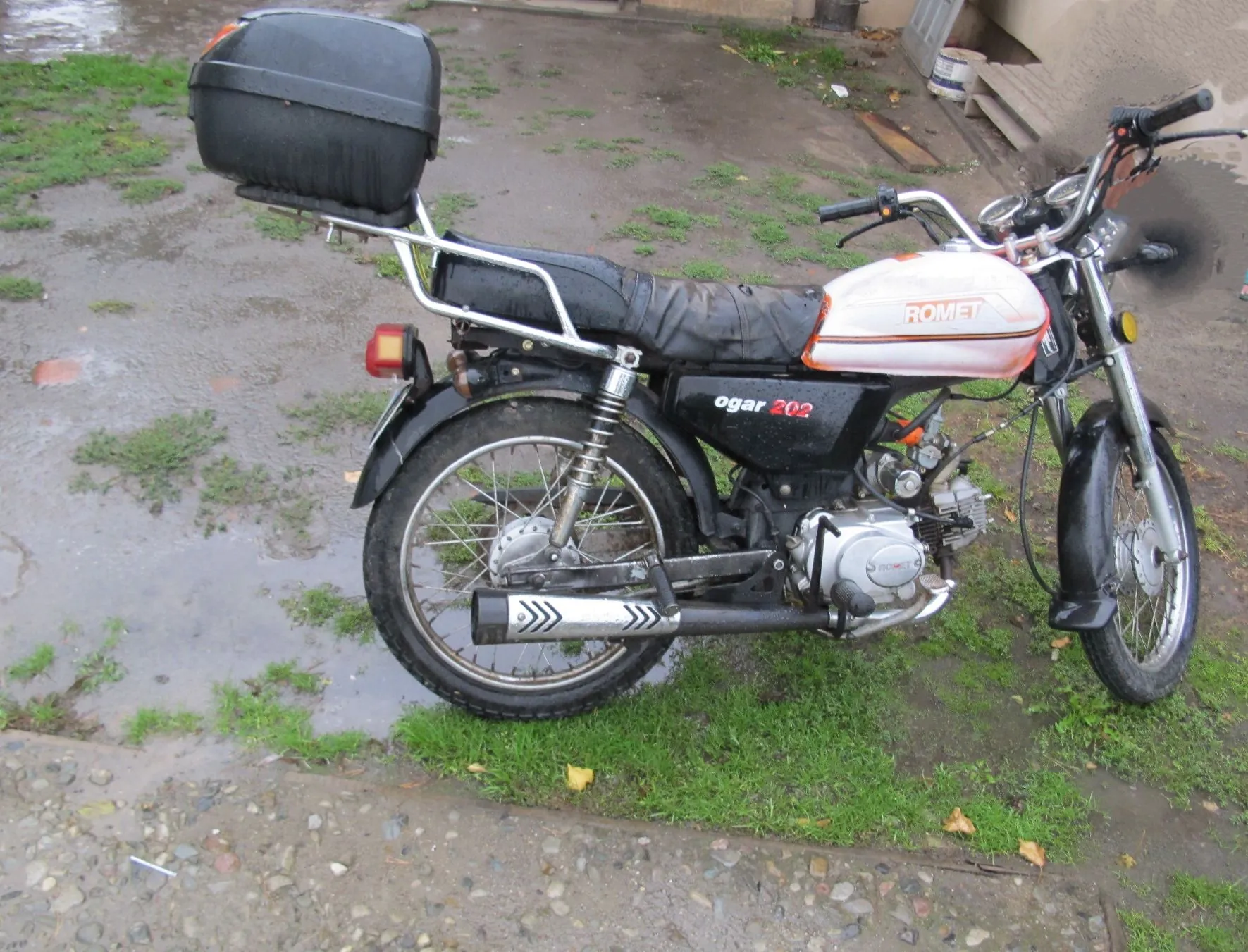 Widok z boku na motocykl marki Romet, model Ogar 202, zaparkowany na mokrym podłożu z trawą. Motocykl ma biały bak z czerwonym napisem ROMET, czarne siedzenie oraz czarny kufer zamontowany na tylnym stelażu. Na boku ramy widnieje napis 'ogar 202'. Pojazd posiada szprychowe koła i srebrny wydech.