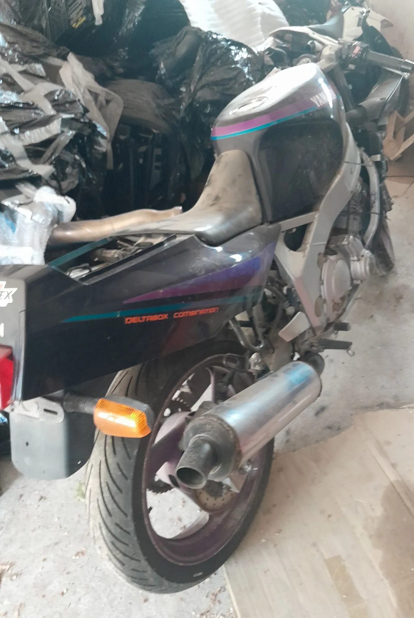 Zdjęcie tylnej części motocykla marki Yamaha w kolorze czarnym z fioletowymi i niebieskimi akcentami. Na boku motocykla widnieje napis 'DELTABOX COMBINATION'. Widoczny jest tylny tłumik wydechowy, koło z oponą oraz pomarańczowy kierunkowskaz. Motocykl znajduje się w pomieszczeniu, w tle widoczne są przedmioty przykryte czarną folią.