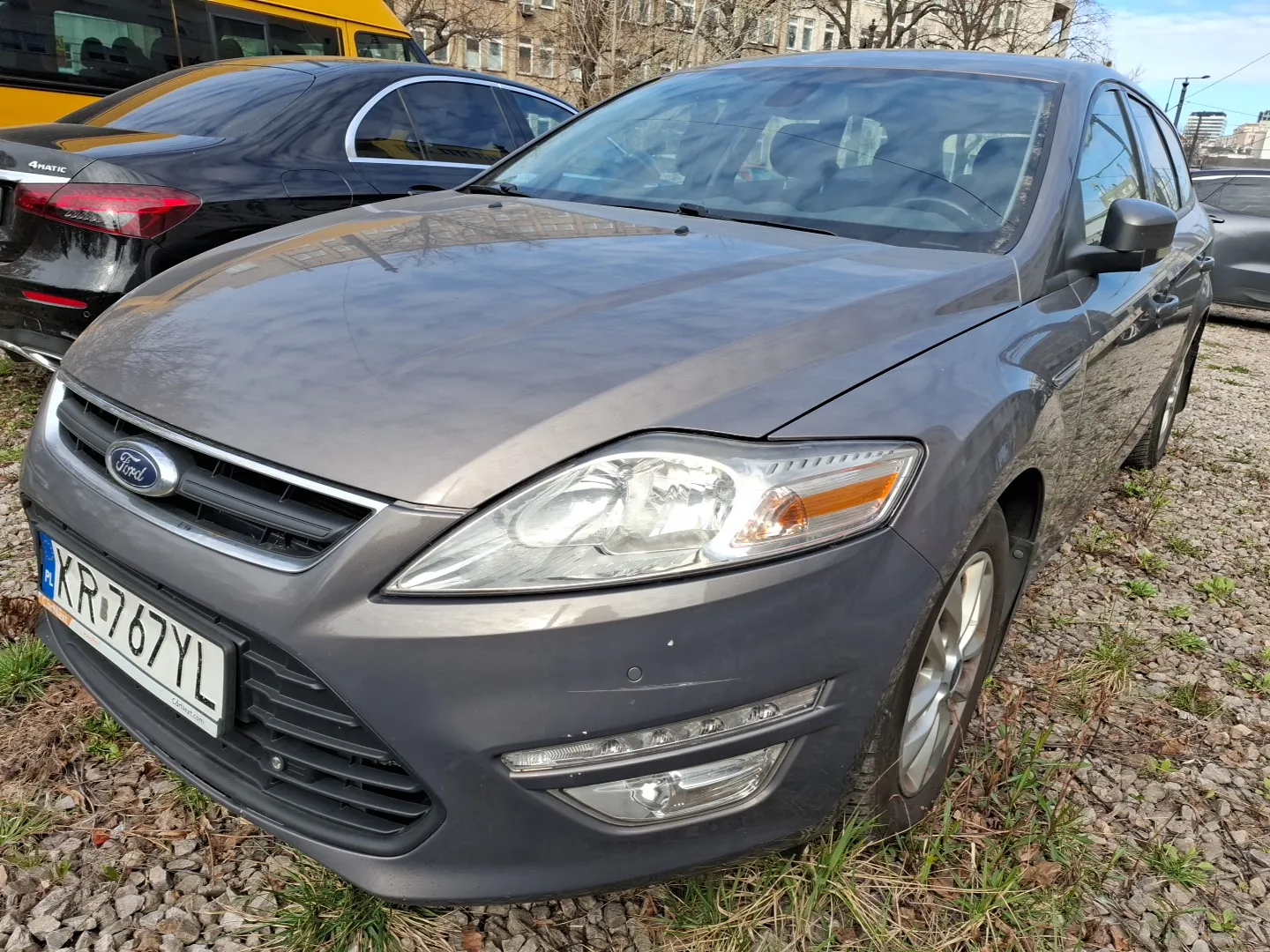 Szary samochód marki Ford z tablicą rejestracyjną KR767YL (PL) zaparkowany na podłożu z żwiru i trawy. W tle czarny samochód z napisem '4MATIC' oraz żółty autobus. Widoczne budynki i drzewa bez liści.