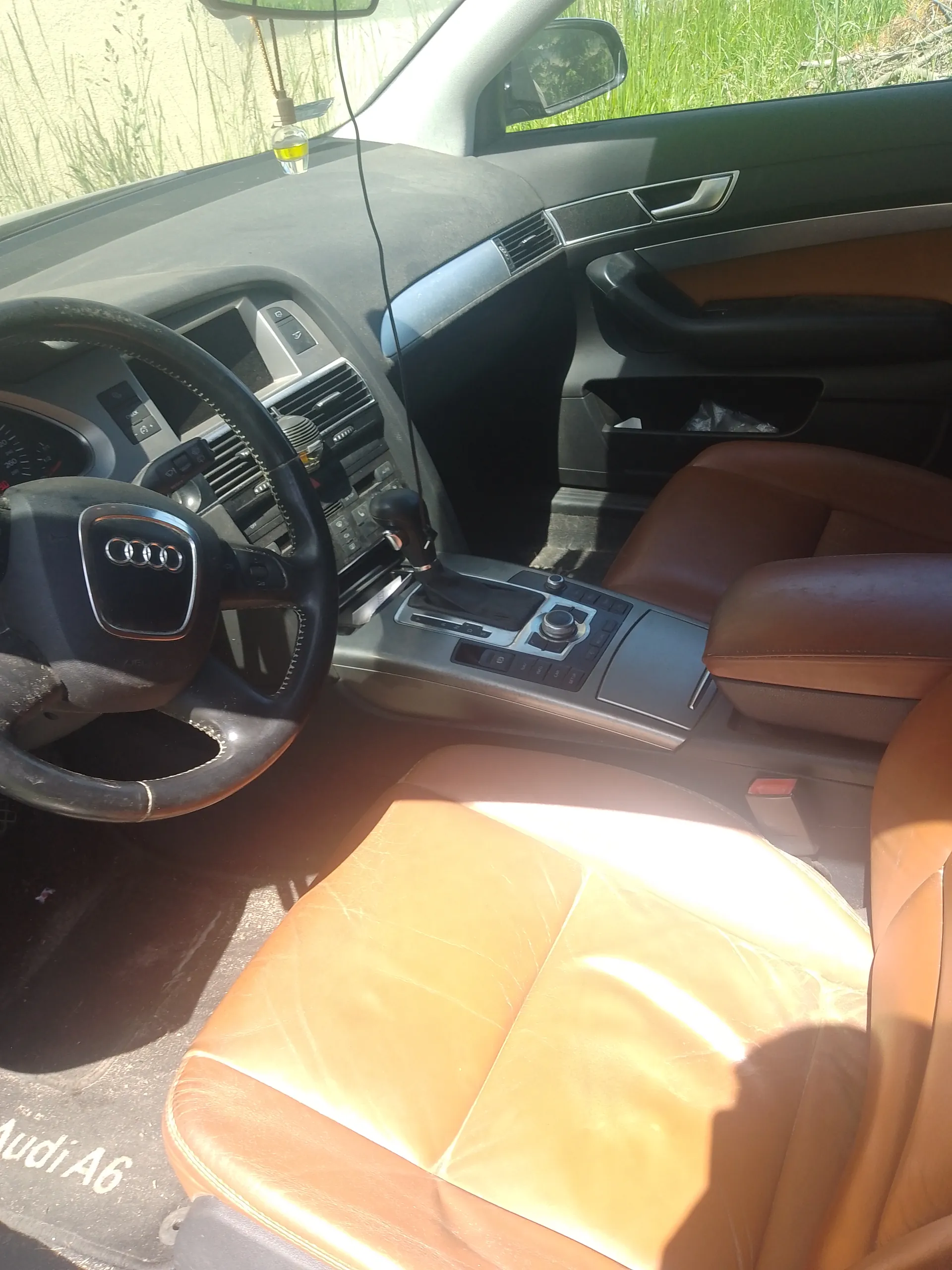 Wnętrze samochodu Audi A6 z widocznym kierownicą z logo Audi (cztery pierścienie), brązowymi skórzany siedzeniami, centralną konsolą z przyciskami i dźwignią zmiany biegów. Na desce rozdzielczej znajdują się wentylacyjne otwory i elementy sterujące. W tle przez przednią szybę widać trawę i ścianę. Na dywaniku pod kierowcą widoczny napis "Audi A6". W lusterku wstecznym zawieszony jest mały klosz z płynem.