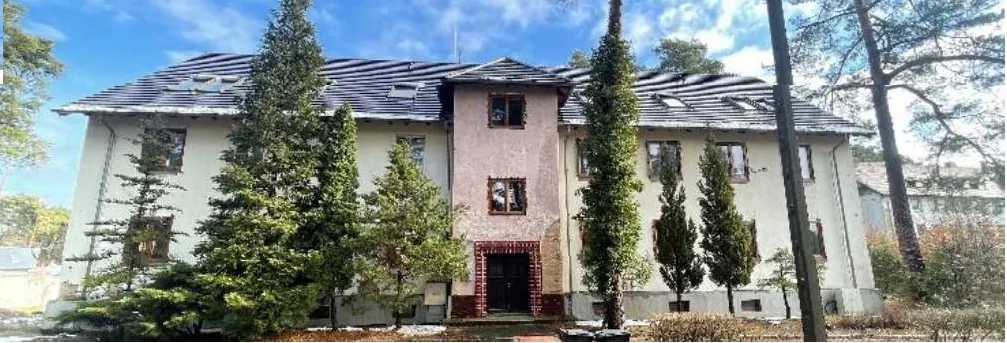 Duży, dwupiętrowy budynek o beżowo-różowej elewacji z ciemnym dachem krytym płytkami, część z nich uszkodzona. W centralnej części budynku czerwona cegła nad wejściem, okna z rozbitymi szybami. Przed budynkiem wysokie, wąskie drzewa, na ziemi śnieg. Tło: błękitne niebo z chmurami, po bokach drzewa i inne budynki. Budynek wydaje się opuszczony lub niedbany.