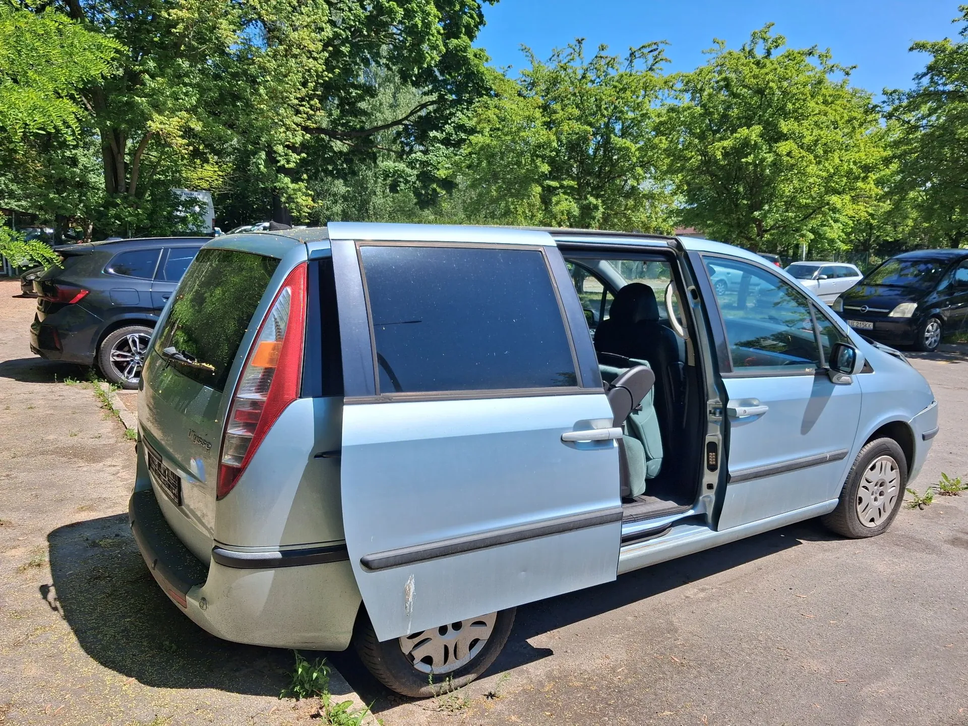 Jasnoniebieski samochód typu minivan zaparkowany na asfaltowym podłożu. Przednie prawe drzwi są otwarte. Samochód ma widoczne uszkodzenia na tylnym zderzaku oraz na dolnej części drzwi pasażera. W tle widać inne zaparkowane samochody oraz gęste, zielone drzewa pod bezchmurnym, niebieskim niebem.