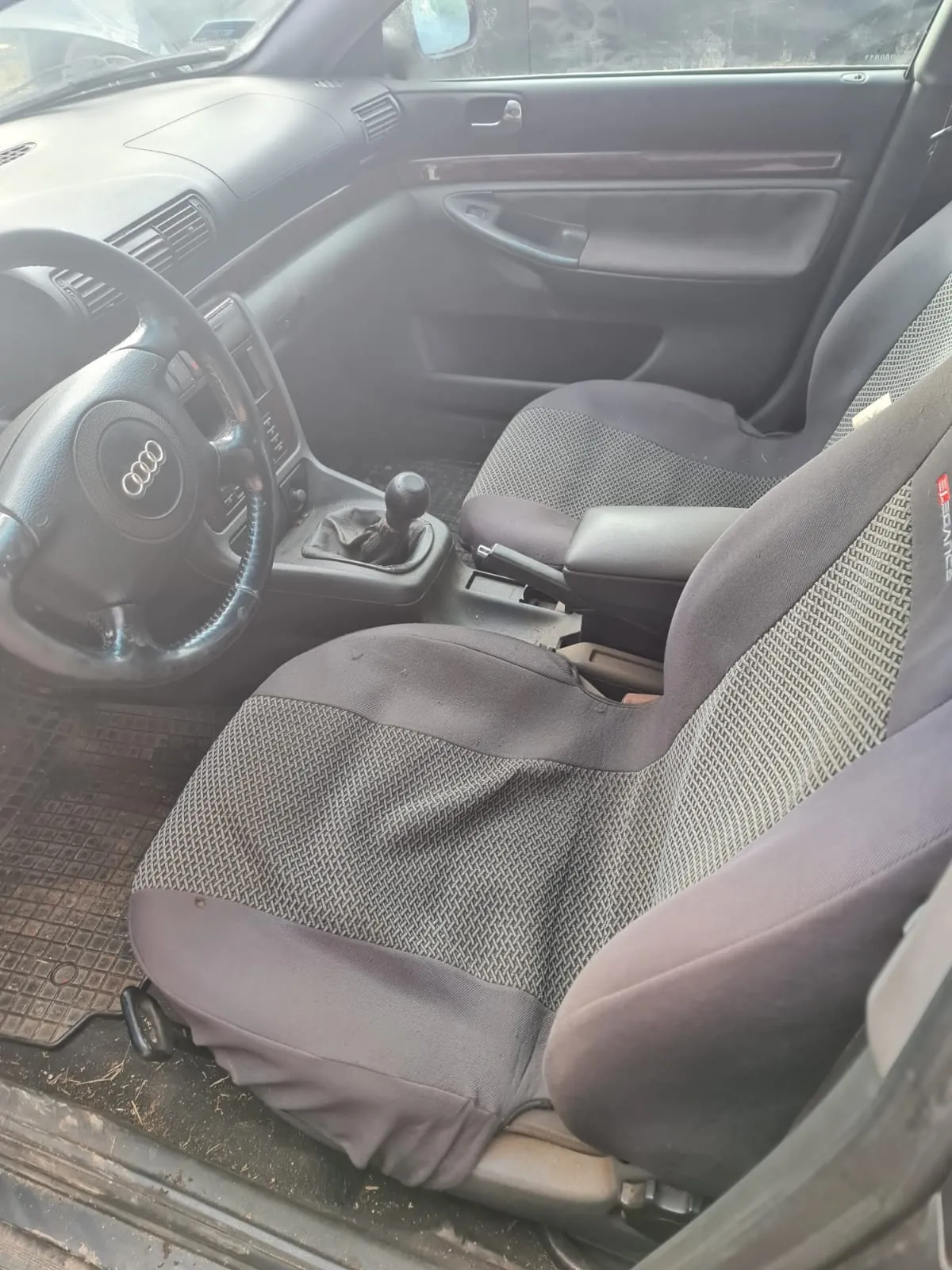 Wnętrze samochodu marki Audi z widocznym kierownicą z logo czterech pierścieni, manualną skrzynią biegów, siedzeniami w kolorze szaro-czarnym z geometrycznym wzorem, deska rozdzielcza z wentylacjami i przyciskami, drzwiami pasażera z uchwytem i okienkami. Podłoga pokryta jest gumową wykładziną z widocznymi śladami brudu. Wnętrze wydaje się nieco zużyte.