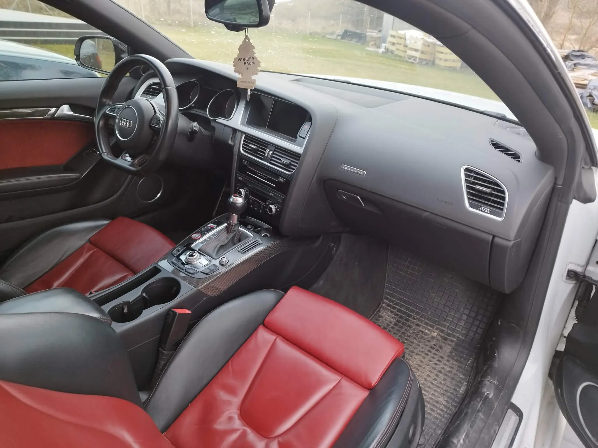 Wnętrze samochodu marki Audi z perspektywy otwartych drzwi pasażera. Widoczna jest czarno-czerwona skórzana tapicerka foteli, czarna deska rozdzielcza, kierownica z logo Audi oraz konsola centralna z lewarkiem zmiany biegów. Na lusterku wstecznym wisi zapach samochodowy w kształcie choinki z napisem 'WUNDER-BAUM'. Podłoga pasażera jest wyłożona gumową matą.