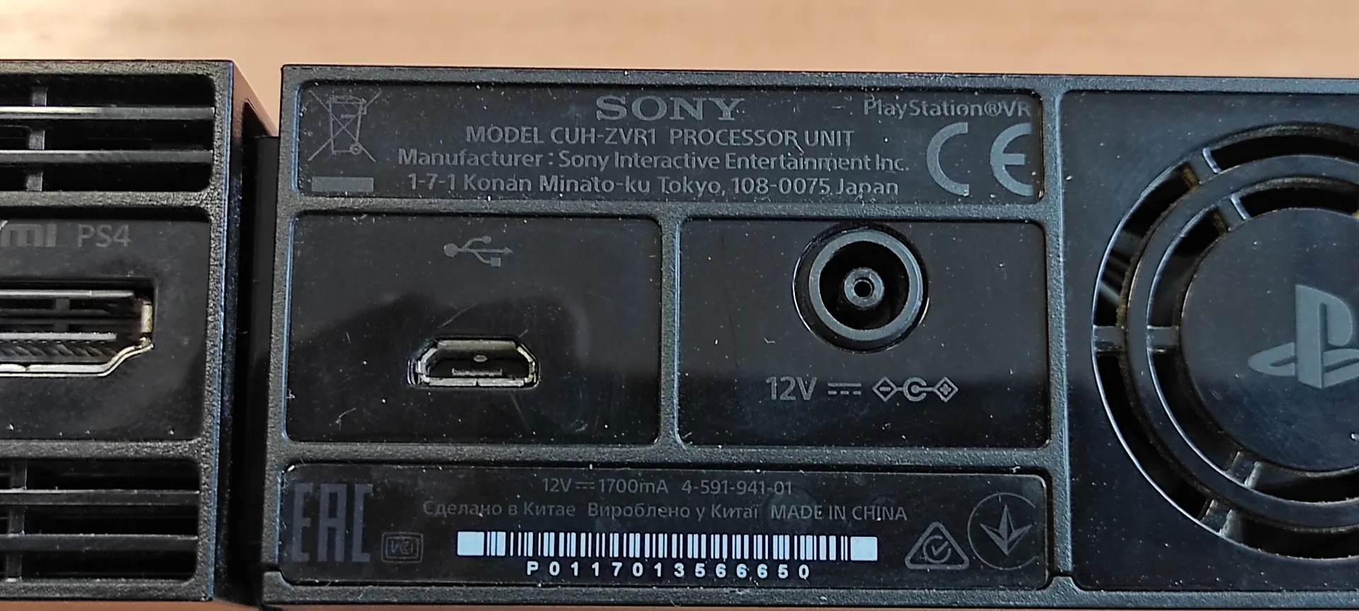 Zbliżenie na tylny panel urządzenia Sony PlayStation VR Processor Unit. Widoczna jest tabliczka znamionowa z tekstem: 'SONY PlayStation VR MODEL CUH-ZVR1 PROCESSOR UNIT Manufacturer: Sony Interactive Entertainment Inc. 1-7-1 Konan Minato-ku Tokyo, 108-0075 Japan'. Na panelu znajdują się gniazda: port USB, gniazdo HDMI oraz gniazdo zasilania oznaczone jako '12V'. Na dole widnieje napis 'MADE IN CHINA' oraz kod kreskowy z numerem 'P0117013566650'. Po prawej stronie znajduje się czarny wentylator z logo PlayStation.