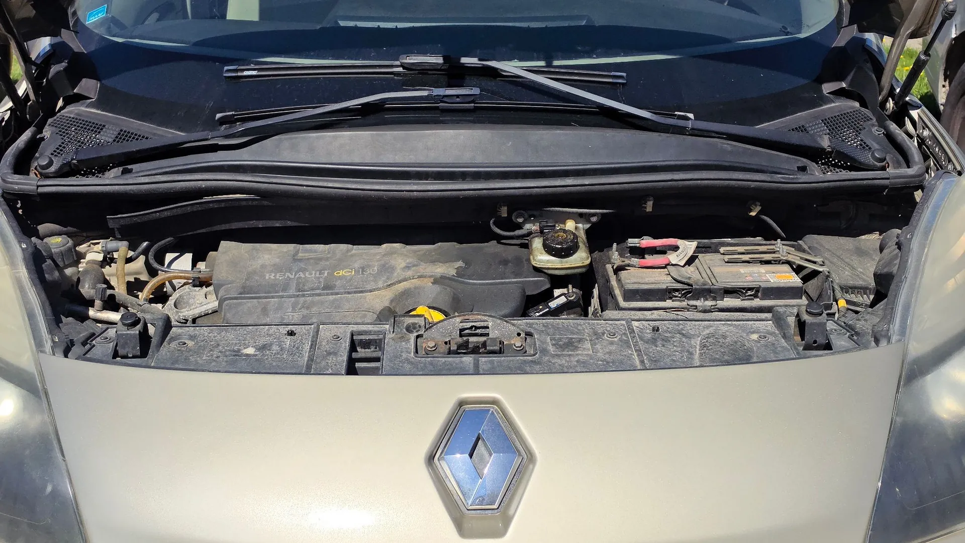Widok z góry na otwartą maskę samochodu marki Renault. Widoczna jest komora silnika z plastikową osłoną z napisem 'RENAULT dCi 130', akumulatorem po prawej stronie oraz zbiornikiem płynu hamulcowego. Na przednim pasie samochodu znajduje się srebrne logo Renault w kształcie rombu.