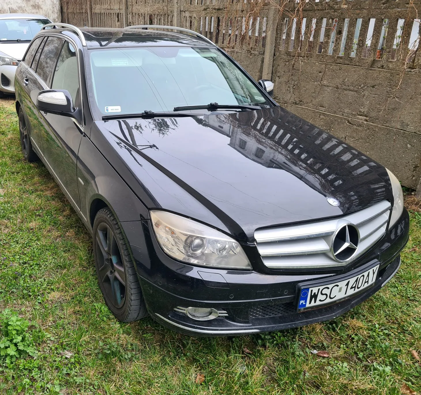Czarny samochód osobowy typu kombi marki Mercedes-Benz zaparkowany na trawie. Widoczna tablica rejestracyjna z napisem PL WSC 140AY. W tle betonowa ściana i drewniane ogrodzenie. Po lewej stronie częściowo widoczny biały samochód. Na przedniej szybie naklejka. Trawa zielona z brązowymi plamami.