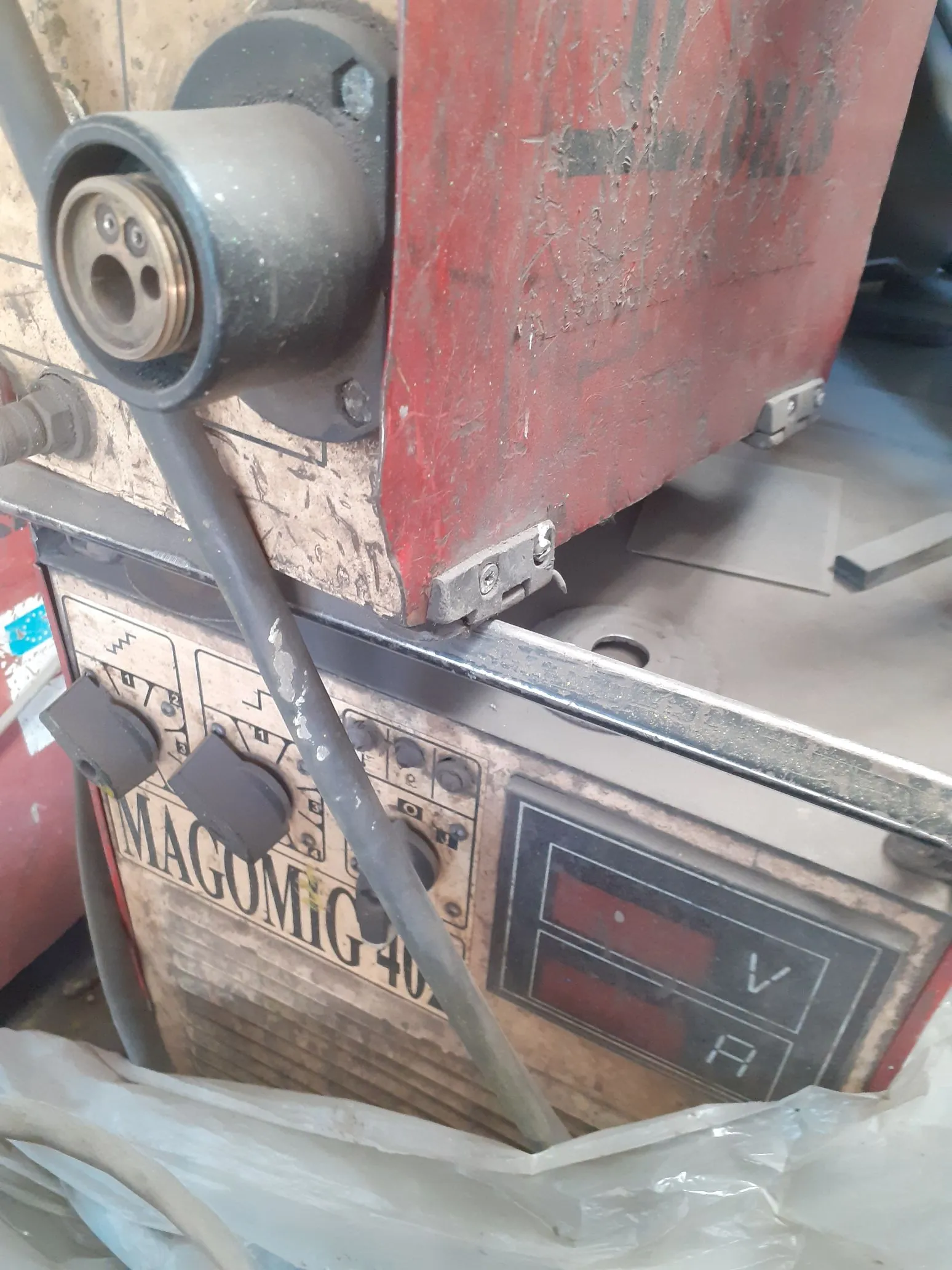 Zbliżenie na fragment czerwonej maszyny przemysłowej, prawdopodobnie spawarki. Widoczny jest panel sterowania z napisem 'MAGOMIG 40', kilkoma pokrętłami oraz wyświetlaczem cyfrowym z oznaczeniami 'V' (wolty) i 'A' (ampery). Do urządzenia podłączony jest gruby, szary kabel.