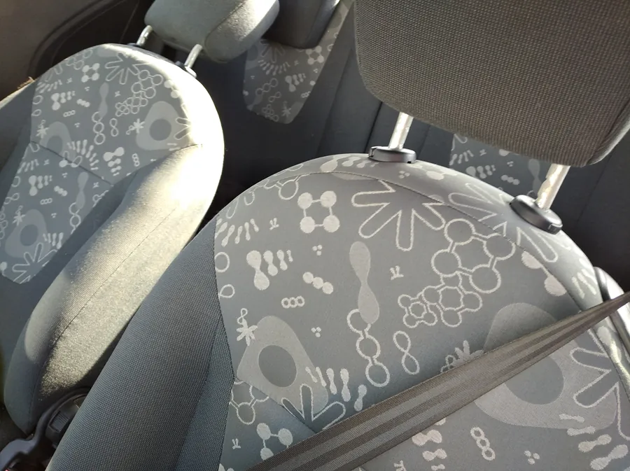 Wnętrze samochodu z widocznymi przednimi siedzeniami pokrytymi tapicerką w szaro-białym wzorze geometrycznym i abstrakcyjnym. Siedzenia mają poduszki z podobnym wzorem, a na jednym z nich widoczny jest pas bezpieczeństwa. W tle częściowo widoczne są poduszki zagłówkowe i fragmenty czarnego wykończenia wnętrza pojazdu.