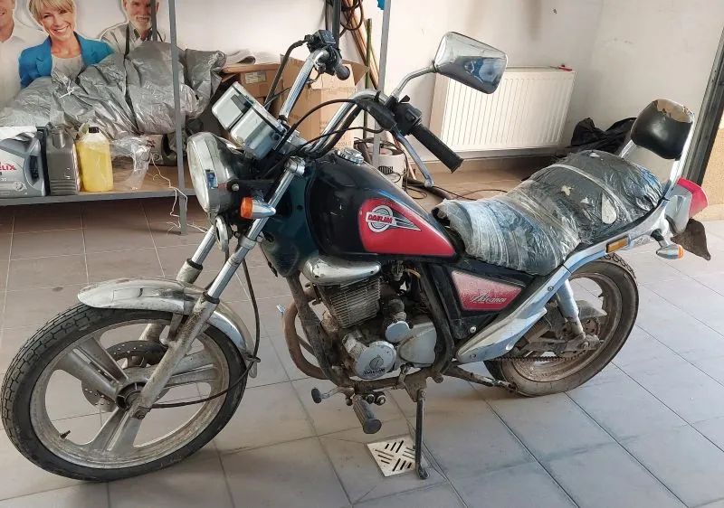 Motocykl marki Honda Cub w kolorze czarno-czerwonym, częściowo przykryty folią, stojący na podpórce w garażu. W tle półka z przedmiotami: żółta kanistra, srebrne worki, narzędzia oraz plakat z dwiema osobami. Podłoga z kafelkami, biała kaloryfer na ścianie. Widoczne elementy motocykla: reflektor, lusterka, tablica rejestracyjna, opony z widocznym brudem.
