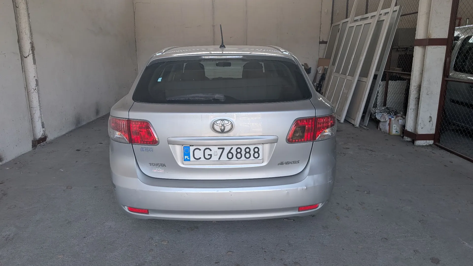Srebrny samochód marki Toyota Avensis zaparkowany tyłem w garażu. Na klapie bagażnika znajduje się logo Toyoty oraz polska tablica rejestracyjna o numerze CG 76888. Po prawej stronie widoczne są oparte o ścianę ramy okienne lub drzwiowe.