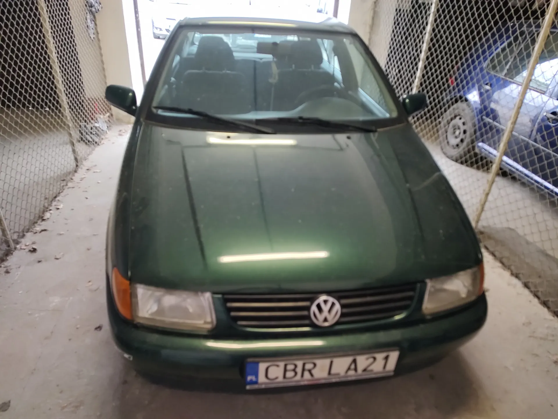 Zielony samochód marki Volkswagen zaparkowany w pomieszczeniu z siatką metalową po obu stronach. Widoczna przód pojazdu z logo VW na atrapie chłodnicy oraz tablicą rejestracyjną CBR LA21 z niebieskim pasem PL po lewej stronie. W tle przez siatkę widoczny niebieski samochód i betonowa podłoga z rozrzuconymi liśćmi.