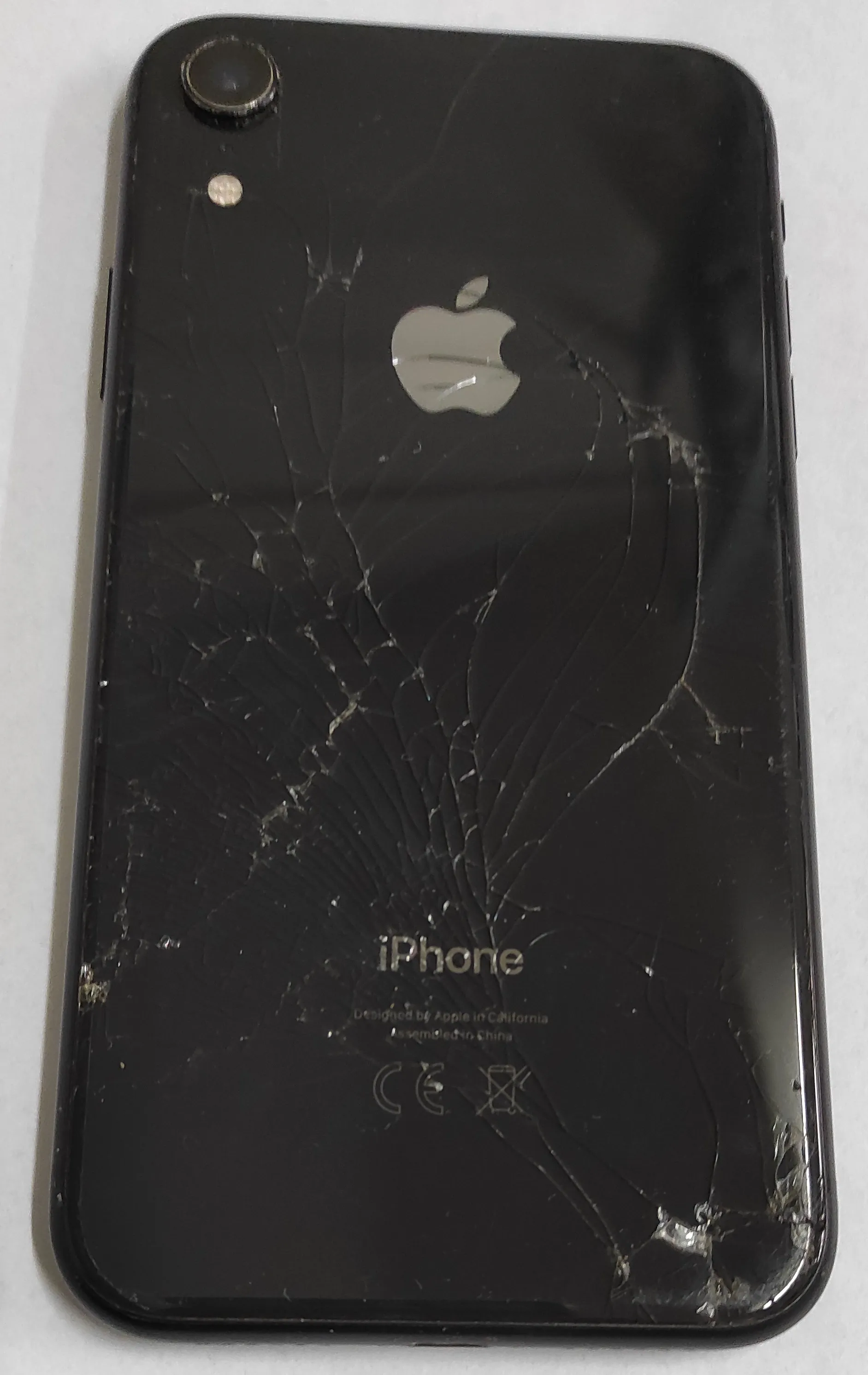 Tylna obudowa czarnego telefonu iPhone z mocno rozbitym szkłem. Na środku znajduje się srebrne logo Apple, a poniżej napis 'iPhone' oraz mniejszy tekst: 'Designed by Apple in California Assembled in China' oraz oznaczenia CE i symbol przekreślonego kosza. W lewym górnym rogu widoczny jest aparat fotograficzny i lampa błyskowa.