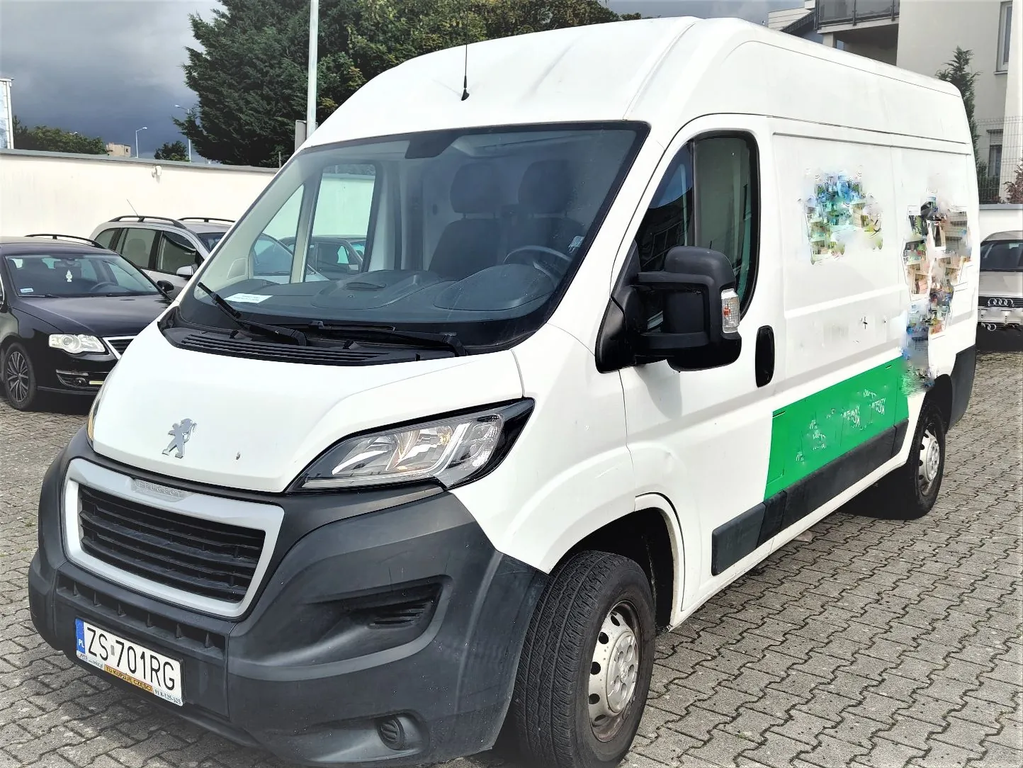 Biały furgon marki Peugeot zaparkowany na kostce brukowej. Pojazd ma czarny zderzak, tablicę rejestracyjną o numerze ZS 701RG oraz zielony pas i zamazane logo na boku nadwozia. W tle widoczne są inne samochody oraz budynek.