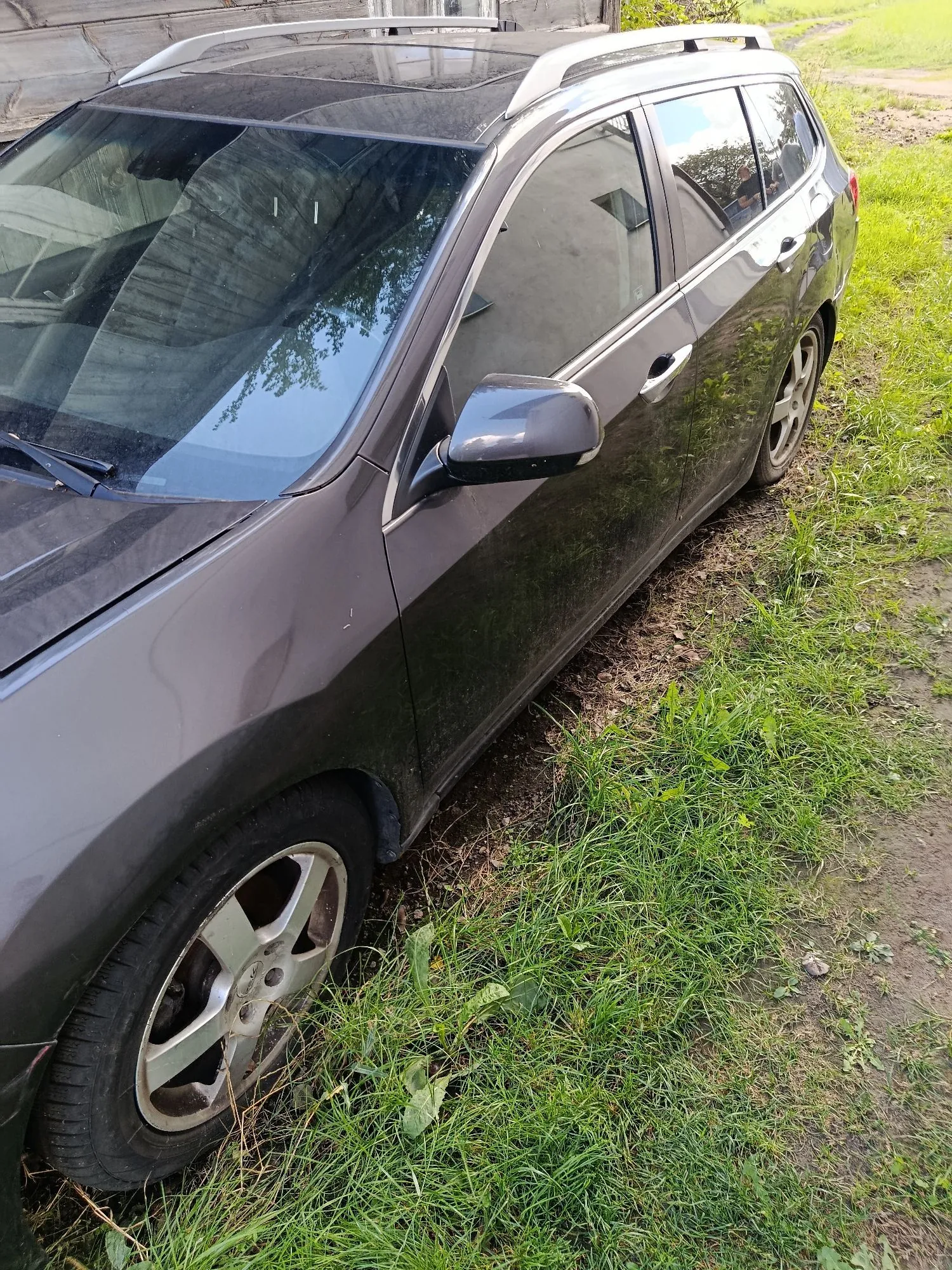 Szary samochód typu kombi zaparkowany na trawiastym poboczu. Widoczny jest bok auta, przednia szyba, lusterko boczne oraz przednie i tylne koło z aluminiowymi felgami. Na dachu samochodu znajdują się srebrne relingi.