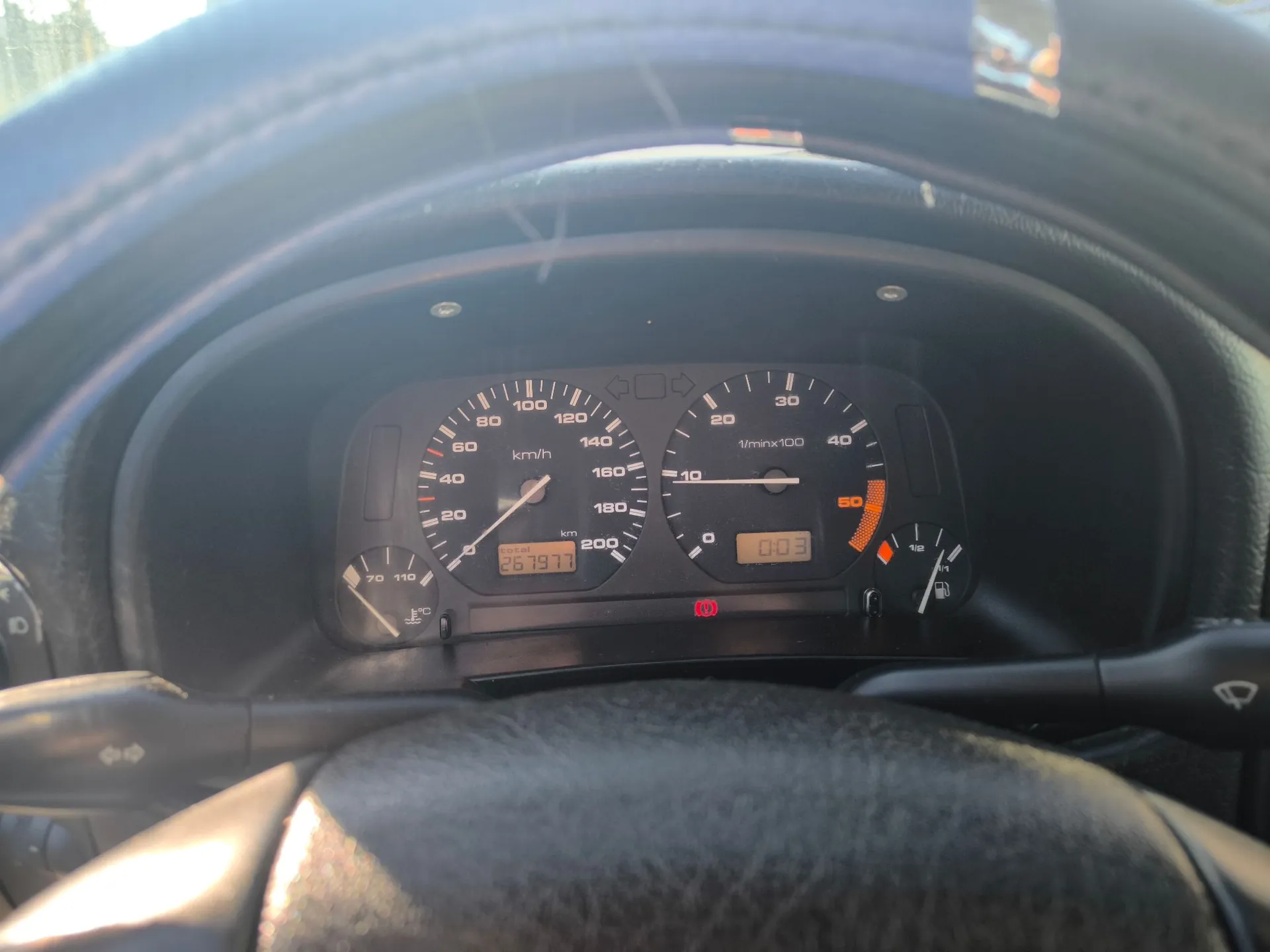 Widok z perspektywy kierowcy na deskę rozdzielczą samochodu. Centralnym elementem jest zestaw wskaźników analogowych: po lewej prędkościomierz z zakresem do 200 km/h, po prawej obrotomierz z zakresem do 40 x 100 1/min. Poniżej prędkościomierza znajduje się licznik kilometrów z wartością 257977 km, a pod obrotomierzem cyfrowy zegar wyświetlający 0:03. Widoczne są również kontrolki ostrzegawcze, w tym czerwona kontrolka z symbolem wykrzyknika w kółku. W kadrze znajduje się fragment kierownicy oraz lewa i prawa wajcha sterująca.