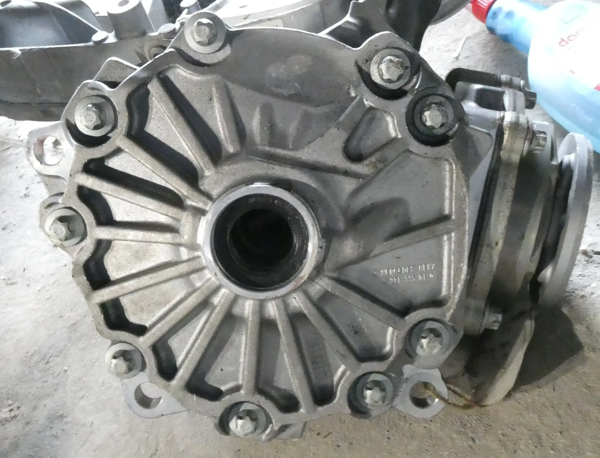 Zbliżenie na metalową, odlewaną część mechaniczną, prawdopodobnie obudowę skrzyni biegów lub dyferencjału. Element ma okrągły kształt z centralnym otworem i promieniście rozmieszczonymi żebrami wzmacniającymi. Obudowa jest skręcona kilkoma śrubami z sześciokątnymi łbami. Na powierzchni metalu widoczne są niewielkie zanieczyszczenia oraz niewyraźne oznaczenia techniczne w postaci numerów i liter.