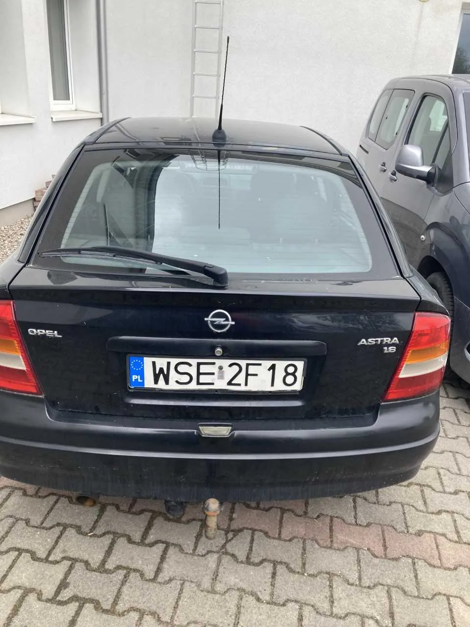Tył czarnego samochodu Opel Astra 1.6 z tablicą rejestracyjną WSE 2F18 (PL) umieszczoną na środku. Po prawej stronie widoczny fragment szarego samochodu. W tle budynek z oknem i metalową drabiną opartą o ścianę. Samochód stoi na brukowanej powierzchni.