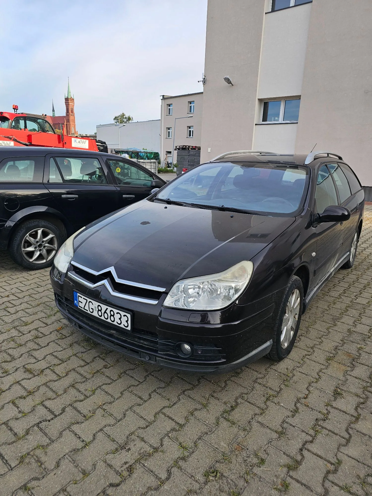Czarny samochód osobowy marki Citroën w nadwoziu kombi zaparkowany na kostce brukowej. Pojazd ma tablicę rejestracyjną o numerze EZG 86833. W tle widoczny jest inny czarny samochód, biały budynek oraz czerwony dźwig i wieża kościoła w oddali.