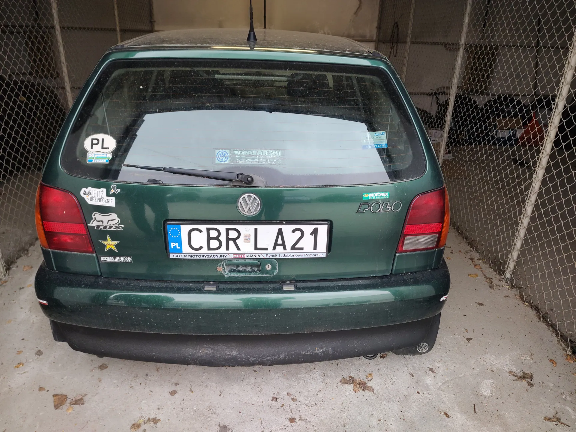 Z tyłu zielony samochód Volkswagen Polo z tablicą rejestracyjną CBR LA21. Na szybie tylnej i nadwoziu widoczne naklejki, w tym okrągła z napisem PL, naklejka z napisem SKLEP MOTORYZACYJNY oraz logo VW w centrum. Samochód zaparkowany na betonowej powierzchni z suchymi liśćmi, w tle siatka ogrodzeniowa i betonowa ściana. Tablica rejestracyjna zawiera również napisy: PL, MOTOKUŹNIA, Rynek 1, Jabłonowo Pomorskie.