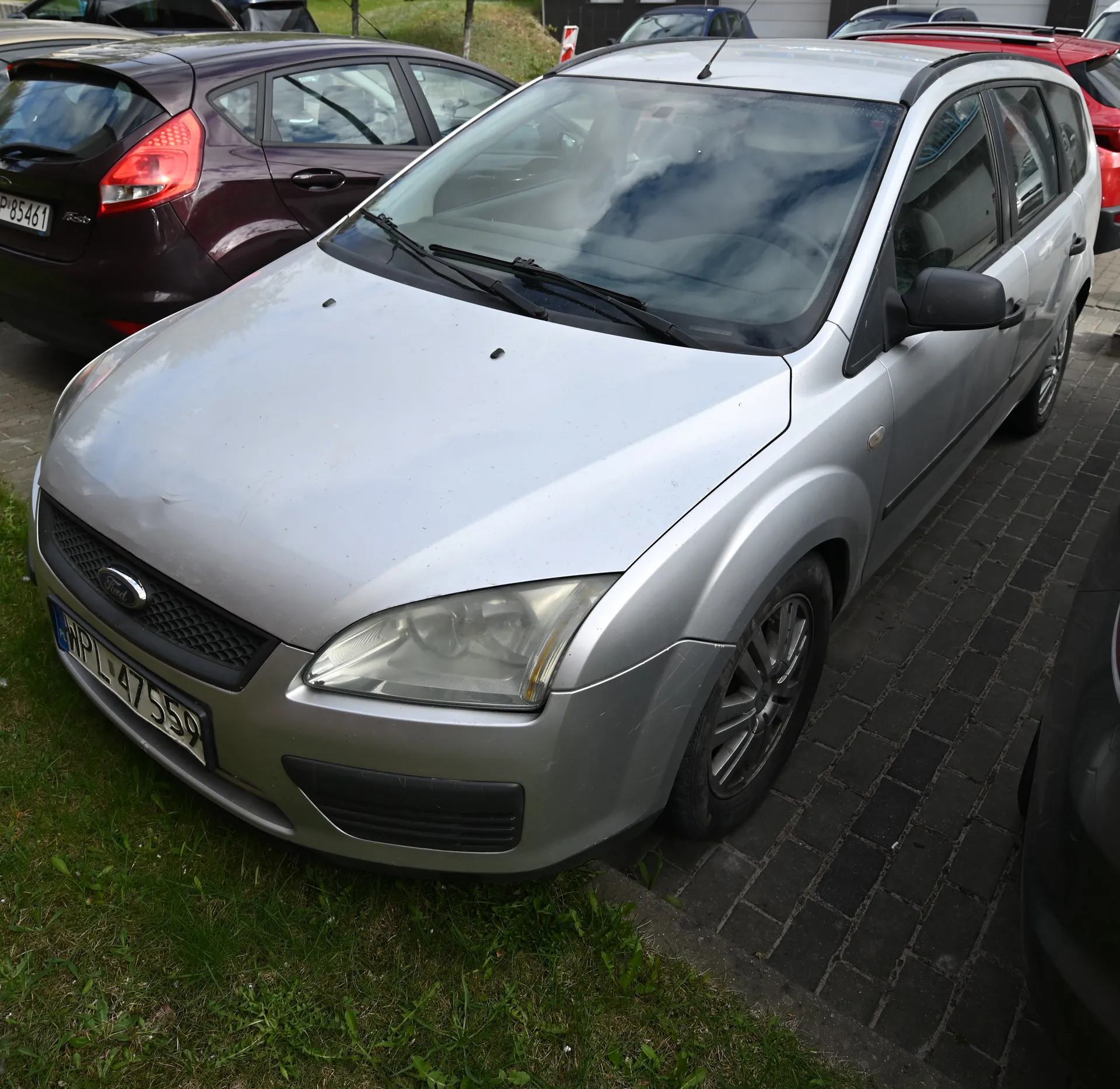 Srebrny samochód marki Ford z tablicą rejestracyjną WPL 47559 zaparkowany na brukowanej powierzchni obok trawnika. W tle widoczne są inne pojazdy, w tym ciemnopurpurowy samochód z tablicą P 85461. Na pierwszym planie trawa, a w oddali fragment budynku i drzewa.