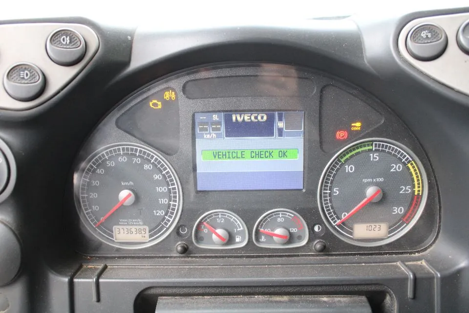 Zbliżenie na deskę rozdzielczą pojazdu marki Iveco. W centrum znajduje się cyfrowy wyświetlacz z napisem 'IVECO' oraz komunikatem 'VEHICLE CHECK OK'. Po lewej stronie widoczny jest prędkościomierz z maksymalną wartością 120 km/h, a po prawej obrotomierz z oznaczeniem 'rpm x100'. Poniżej ekranu znajdują się dwa mniejsze wskaźniki: poziom paliwa oraz temperatura cieczy chłodzącej. Pod prędkościomierzem widnieje licznik przebiegu wskazujący wartość 373638.9. Nad ekranem świecą się żółte kontrolki ostrzegawcze, a po prawej stronie czerwona kontrolka hamulca ręcznego (P).