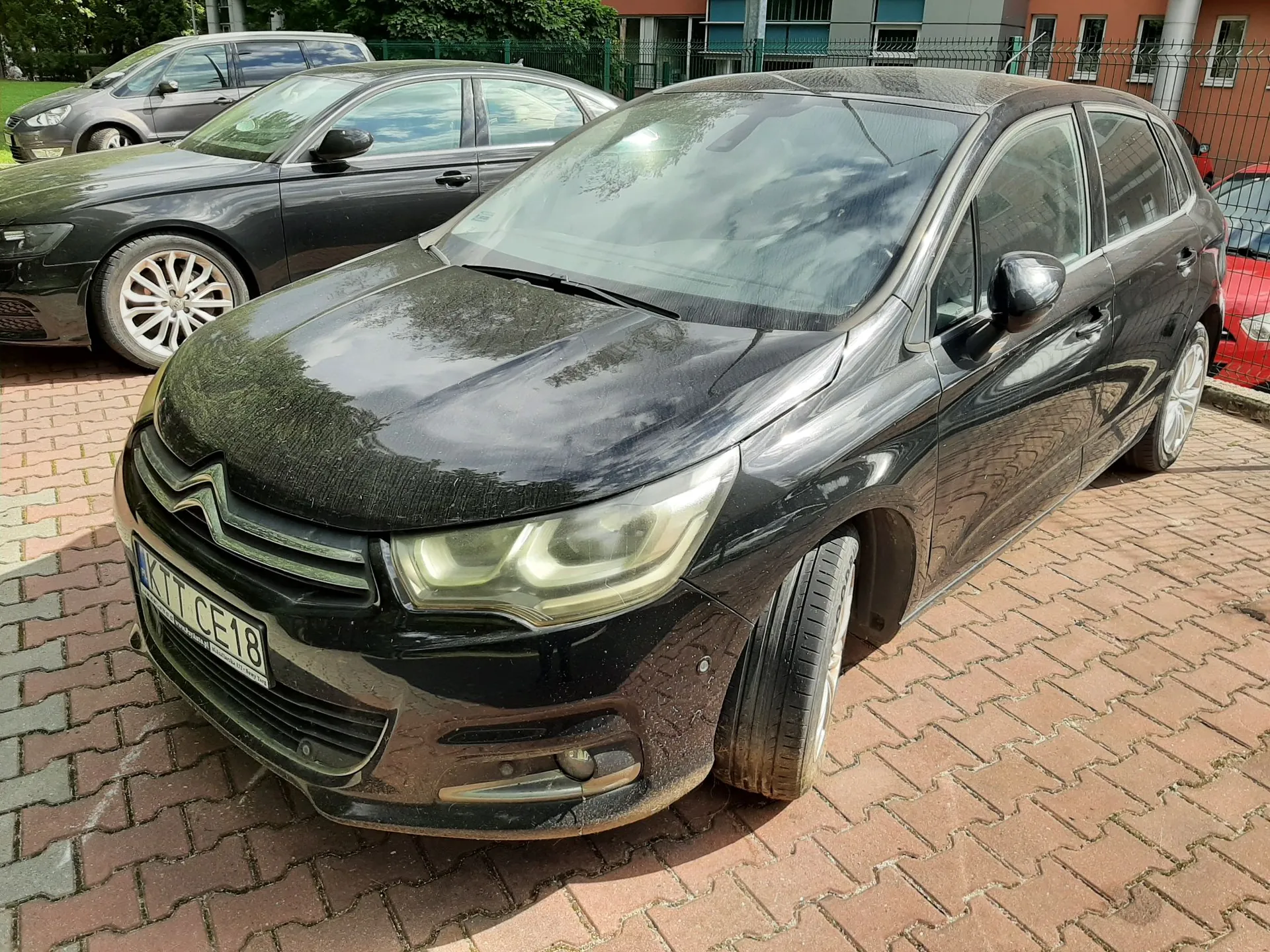 Czarny, zakurzony samochód osobowy marki Citroën zaparkowany na kostce brukowej. W tle widoczny jest inny czarny samochód oraz fragmenty budynków i zieleni. Na przednim zderzaku znajduje się tablica rejestracyjna z fragmentem tekstu 'CE18'.