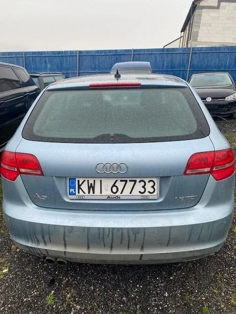 Tył jasnoniebieskiego samochodu marki Audi z tablicą rejestracyjną KW 67733 (PL). Na tle widoczne są inne pojazdy, niebieskie ogrodzenie i budynek. Samochód ma zabrudzenia na tylnym zderzaku. Logo Audi oraz napis Audi umieszczone są pod szybą. W tle po prawej stronie widoczny jest czarny samochód marki Volkswagen.