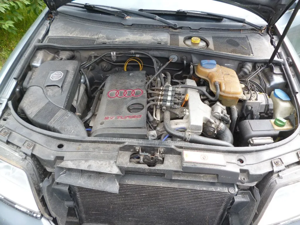 Widok z góry na komorę silnika samochodu marki Audi. W centrum znajduje się silnik z plastikową osłoną, na której widnieją cztery czerwone pierścienie logo Audi oraz napis 'V6 TURBO'. Po prawej stronie widoczny jest żółty zbiornik z płynem chłodniczym, a po lewej czarna obudowa filtra powietrza. Na dole zdjęcia znajduje się chłodnica. Całość jest zakurzona.
