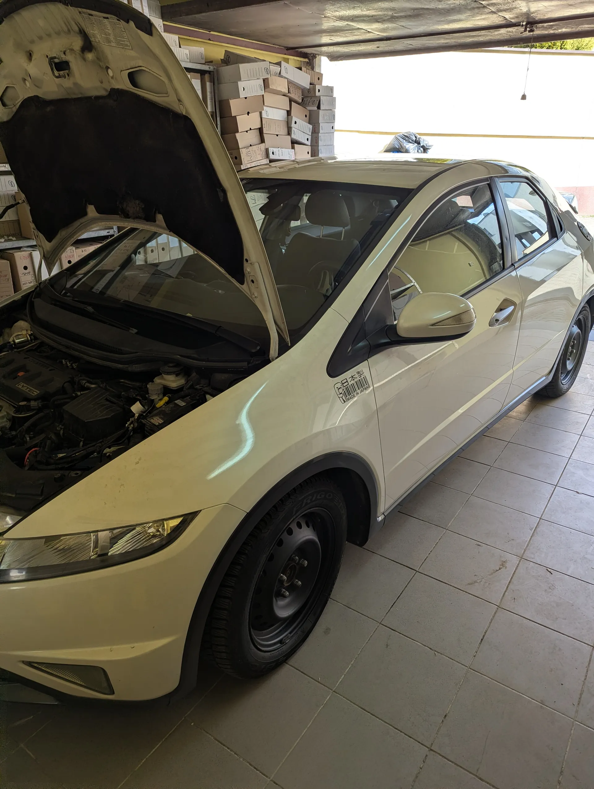 Biały samochód osobowy z otwartą maską ukazującą silnik, zaparkowany w garażu. W tle stosy kartonów. Na drzwiach pasażerskich naklejka z napisem „日本製” oraz kodem kreskowym. Podłoga wyłożona płytkami. Garaż ma dach i otwarte wejście.