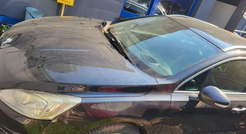 Zbliżenie na przód i przednią szybę czarnego samochodu. Karoseria i szyba są pokryte grubą warstwą kurzu i brudu. Widoczna jest prawa reflektor oraz lusterko boczne.