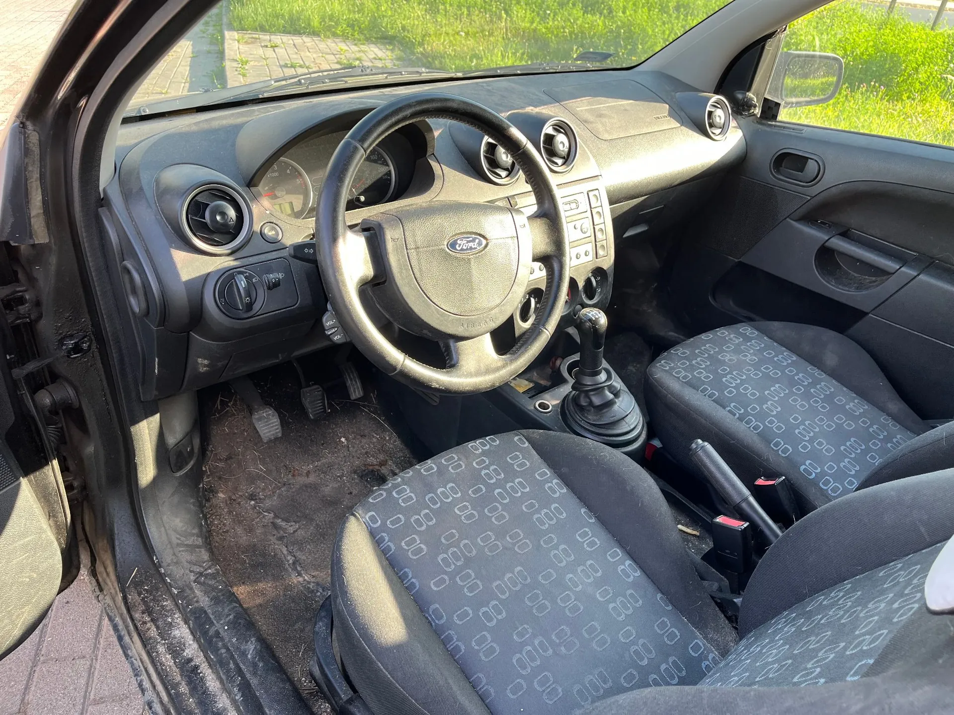 Wnętrze samochodu marki Ford widziane z otwartych drzwi kierowcy. Widoczna jest czarna deska rozdzielcza z licznikiem, kierownica z logo Ford w centrum, otwory nawiewów oraz konsola środkowa z przyciskami. Fotele są obite ciemnoszarą tkaniną w jasny, geometryczny wzór z powtarzającymi się symbolami. Na podłodze widoczne są pedały oraz dywaniki.