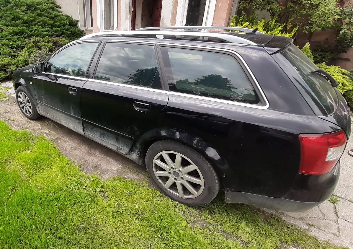 Czarny samochód typu kombi zaparkowany na betonowym podjeździe obok zielonej trawy. Pojazd ma srebrne relingi dachowe oraz wieloramienne felgi aluminiowe. W tle widoczny jest fragment budynku oraz zielone krzewy.