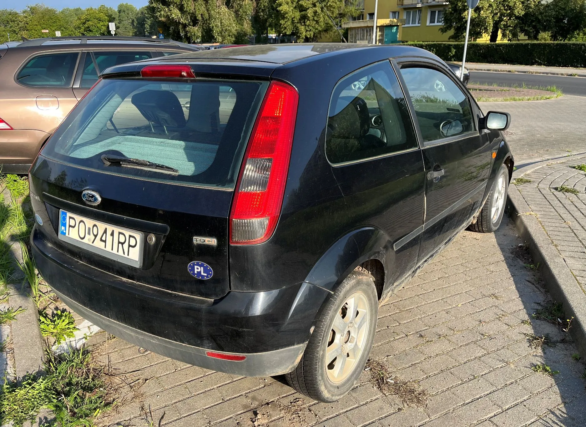 Czarny samochód marki Ford Fiesta zaparkowany tyłem na kostce brukowej. Widoczna jest polska tablica rejestracyjna o numerze PO 941RP oraz naklejka PL. Obok stoi brązowy samochód, a w tle widać zieleń i budynek mieszkalny.