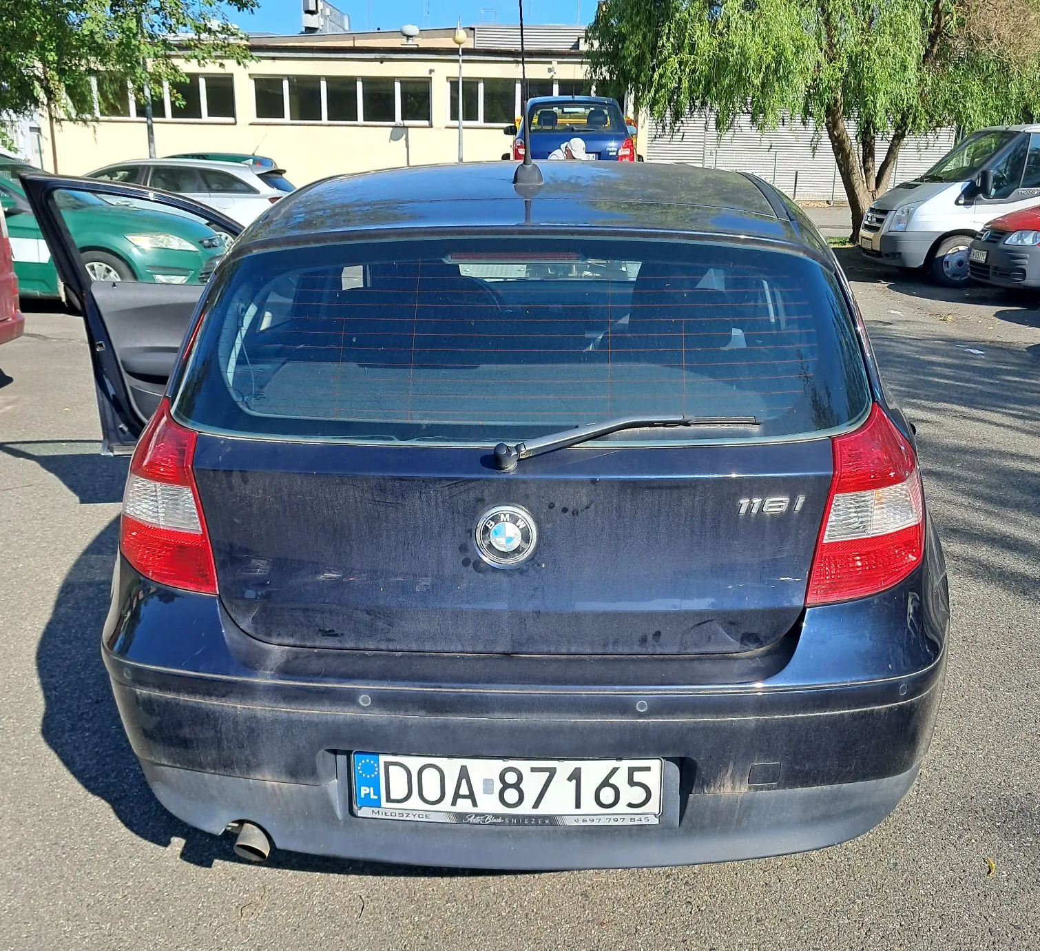 Tylny widok ciemnoniebieskiego samochodu marki BMW serii 1. Pojazd posiada polską tablicę rejestracyjną o numerze DOA 87165. Na klapie bagażnika widoczne jest logo BMW oraz oznaczenie modelu 118i. Samochód zaparkowany jest na asfaltowym placu, w tle widoczne są inne samochody oraz jasny budynek z rzędem okien i zielone drzewa.
