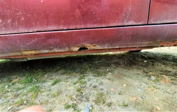 Dolna część czerwonego pojazdu z widoczną rdzą i otworem w metalu. Pod pojazdem ziemia z trawą i drobnymi kamieniami. W lewym dolnym rogu widoczny palec wskazujący w kierunku uszkodzenia.