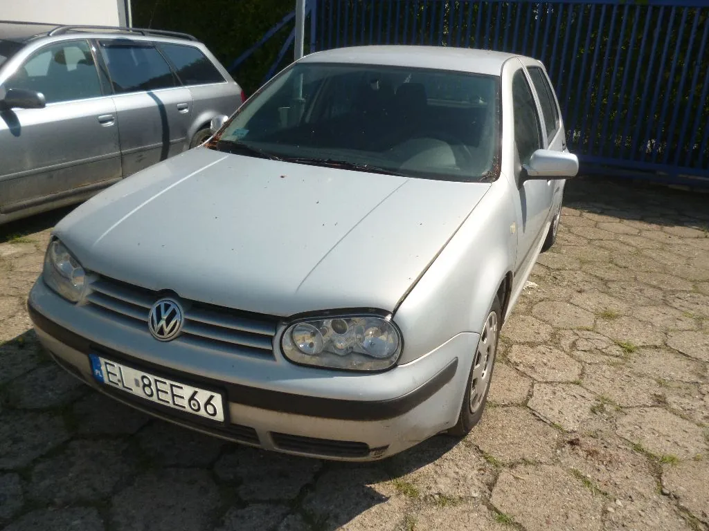 Srebrny samochód osobowy marki Volkswagen Golf zaparkowany na kostce brukowej. Widoczna jest przednia część pojazdu z tablicą rejestracyjną o numerze EL 8EE66. W tle znajduje się inny srebrny samochód oraz niebieski metalowy płot.