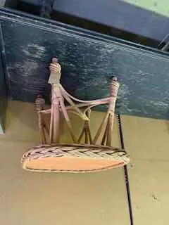 Stare krzesło z plecionym siedziskiem i jasnymi drewnianymi nogami, ustawione obok ciemnego, zużytego stołu. Tło stanowi jasna ściana z czarnym przewodem.