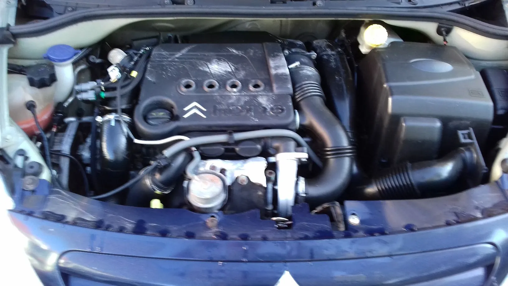 Widok z góry na komorę silnika samochodu marki Citroën. W centrum znajduje się czarna plastikowa osłona silnika z logo Citroën i czterema okrągłymi otworami. Po lewej stronie widoczny jest zbiornik z płynem z niebieskim korkiem, a po prawej czarna obudowa filtra powietrza. Widoczne są liczne przewody, rury dolotowe oraz elementy mechaniczne silnika.