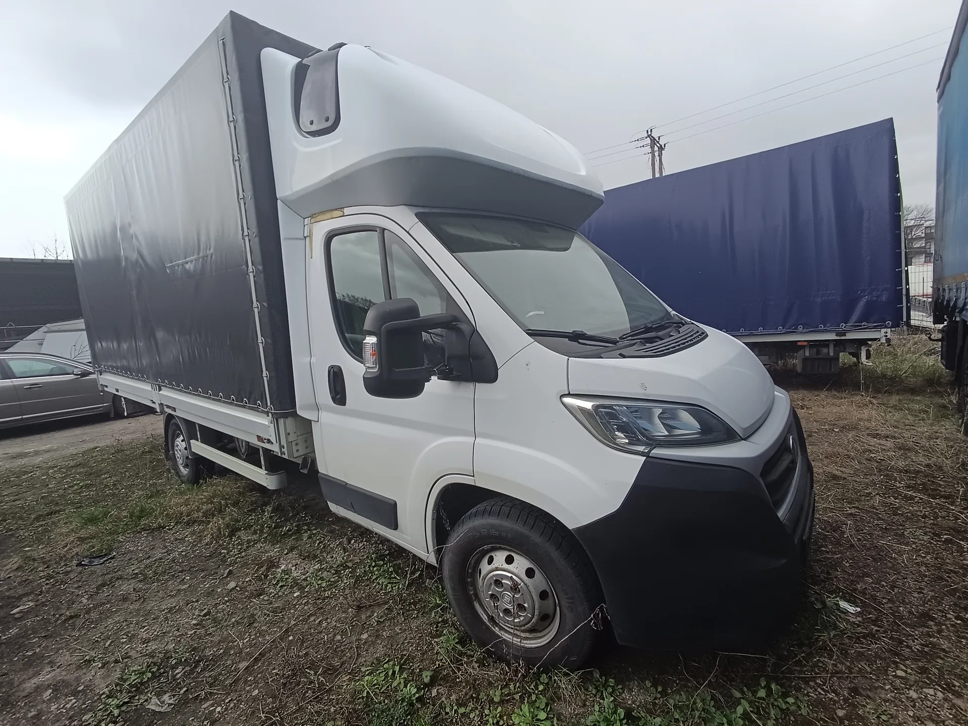 Biały samochód dostawczy z czarnym naczepą na tle otwartego terenu. Po lewej stronie widoczny srebrny samochód osobowy, po prawej niebieska naczepa i fragment niebieskiego pojazdu. Tło stanowi chmurne niebo, słupy energetyczne oraz sucha trawa na ziemi. Samochód dostawczy ma czarne zderzaki, lusterka i opony z metalowymi felgami. W tle również widoczne są inne pojazdy i budynki.