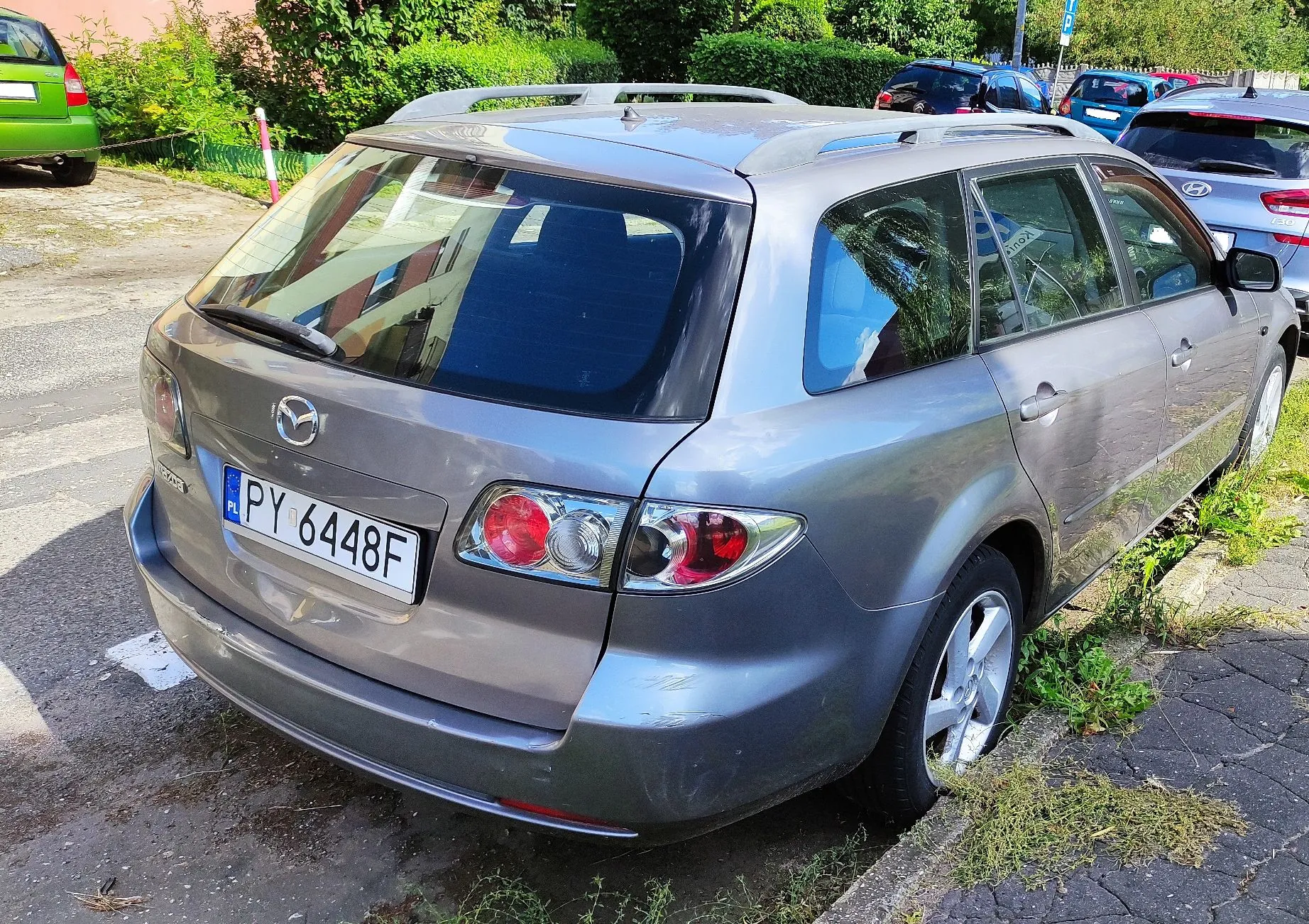 Srebrnoszary samochód marki Mazda w nadwoziu kombi, zaparkowany tyłem do obiektywu na poboczu drogi. Na tylnej klapie widoczna jest polska tablica rejestracyjna o numerze PY 6448F. Samochód posiada relingi dachowe. W tle widoczne są inne zaparkowane pojazdy oraz zielone krzewy i drzewa.