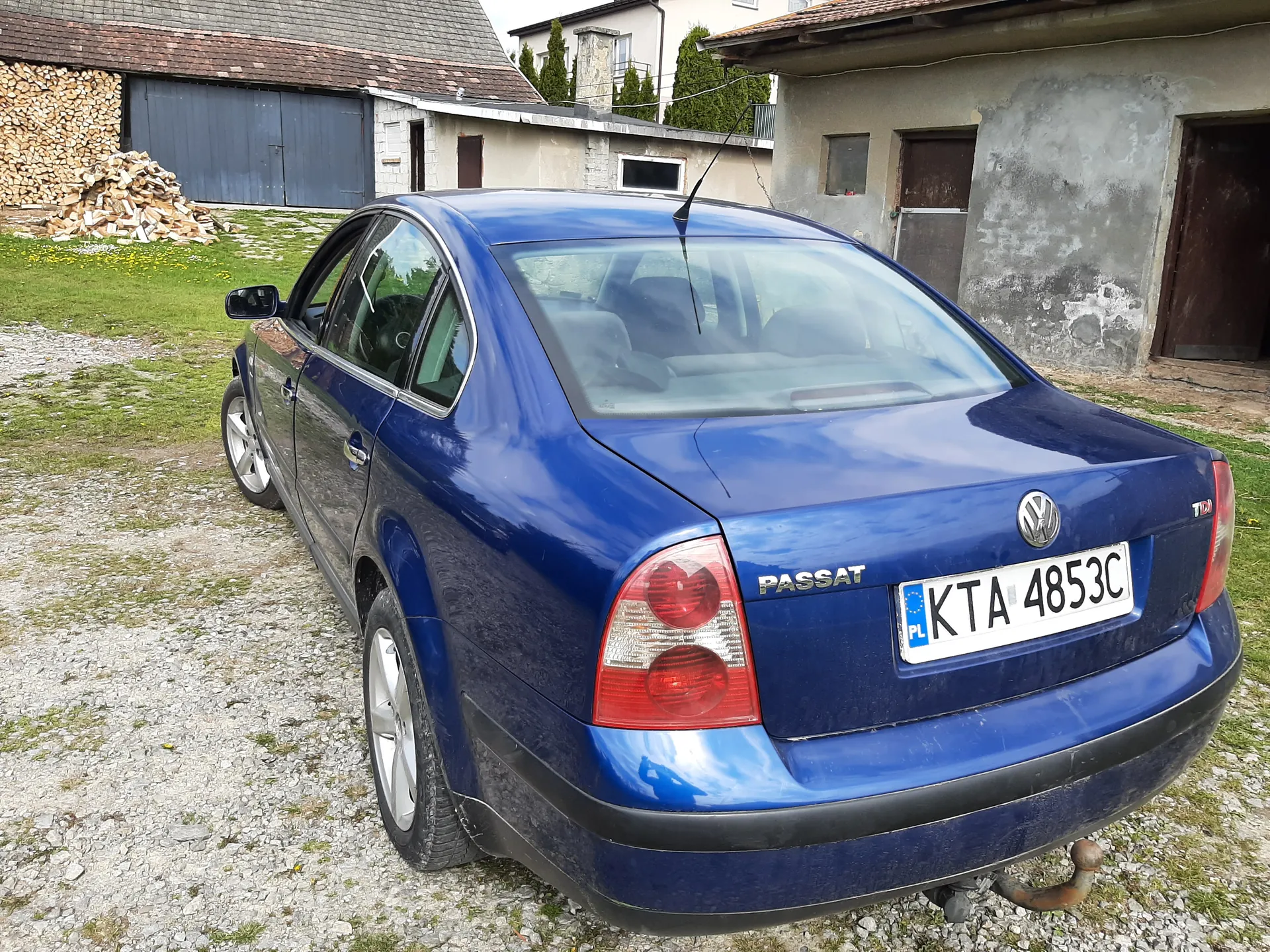 Niebieski samochód osobowy Volkswagen Passat z tablicą rejestracyjną KTA 4853C (PL) zaparkowany na żwirowym podwórku. W tle widoczne są budynki: po lewej stronie dom z niebieskimi drzwiami garażowymi i stosem drewna, po prawej budynek z zatartą elewacją i brązowymi drzwiami. Na podwórku rośnie trawa, a na dachu budynku po lewej stronie znajdują się czerwone dachówki. Samochód ma czerwone światła tylna i widoczny napis PASSAT na bagażniku.