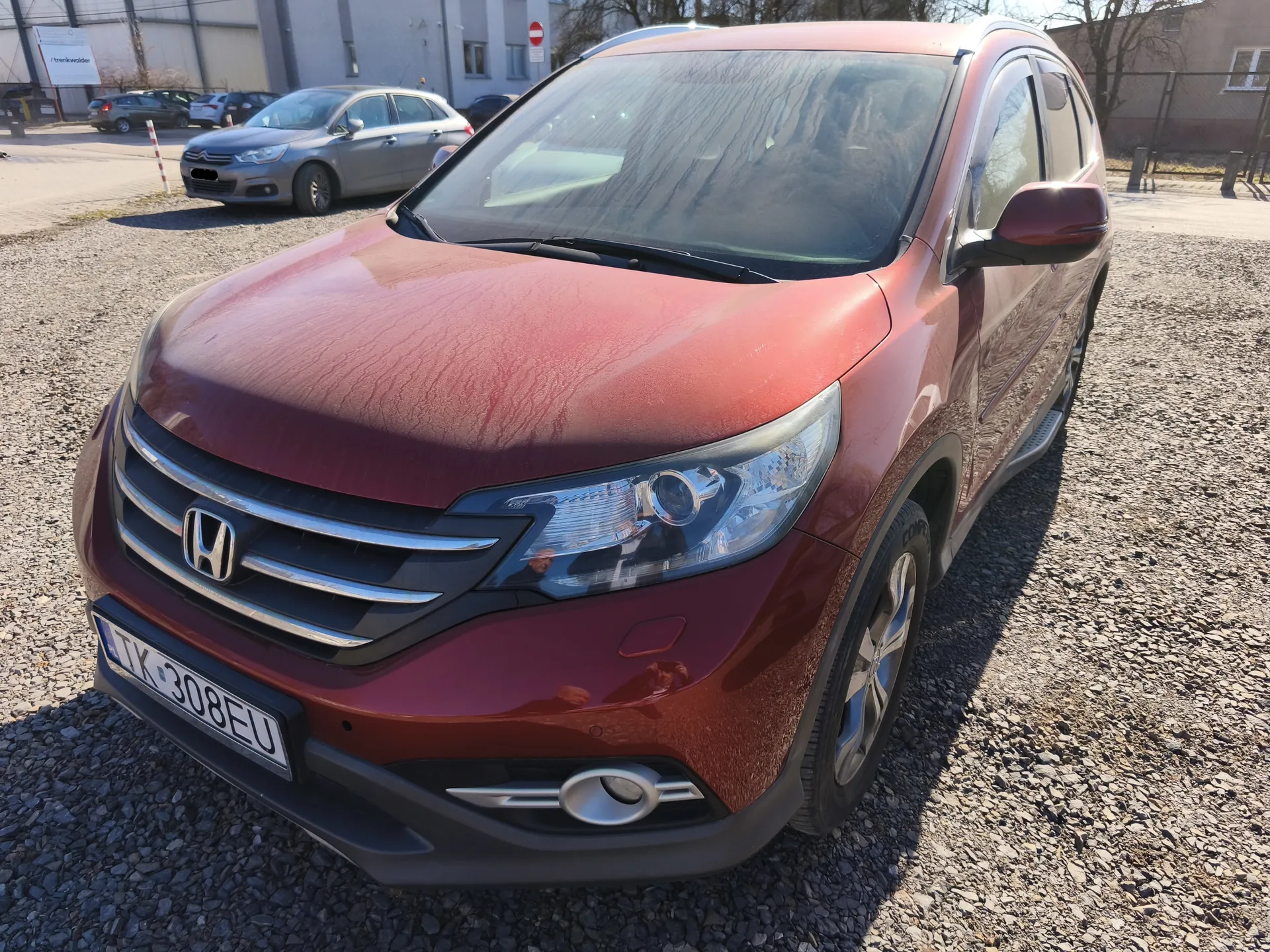 Czerwony samochód osobowy marki Honda CR-V z tablicą rejestracyjną TK 308EU zaparkowany na żwirowym placu. W tle widoczne są inne pojazdy, budynek przemysłowy, znak zakazu wjazdu oraz drzewa. Słoneczna pogoda, cień rzucany przez samochód na żwir.