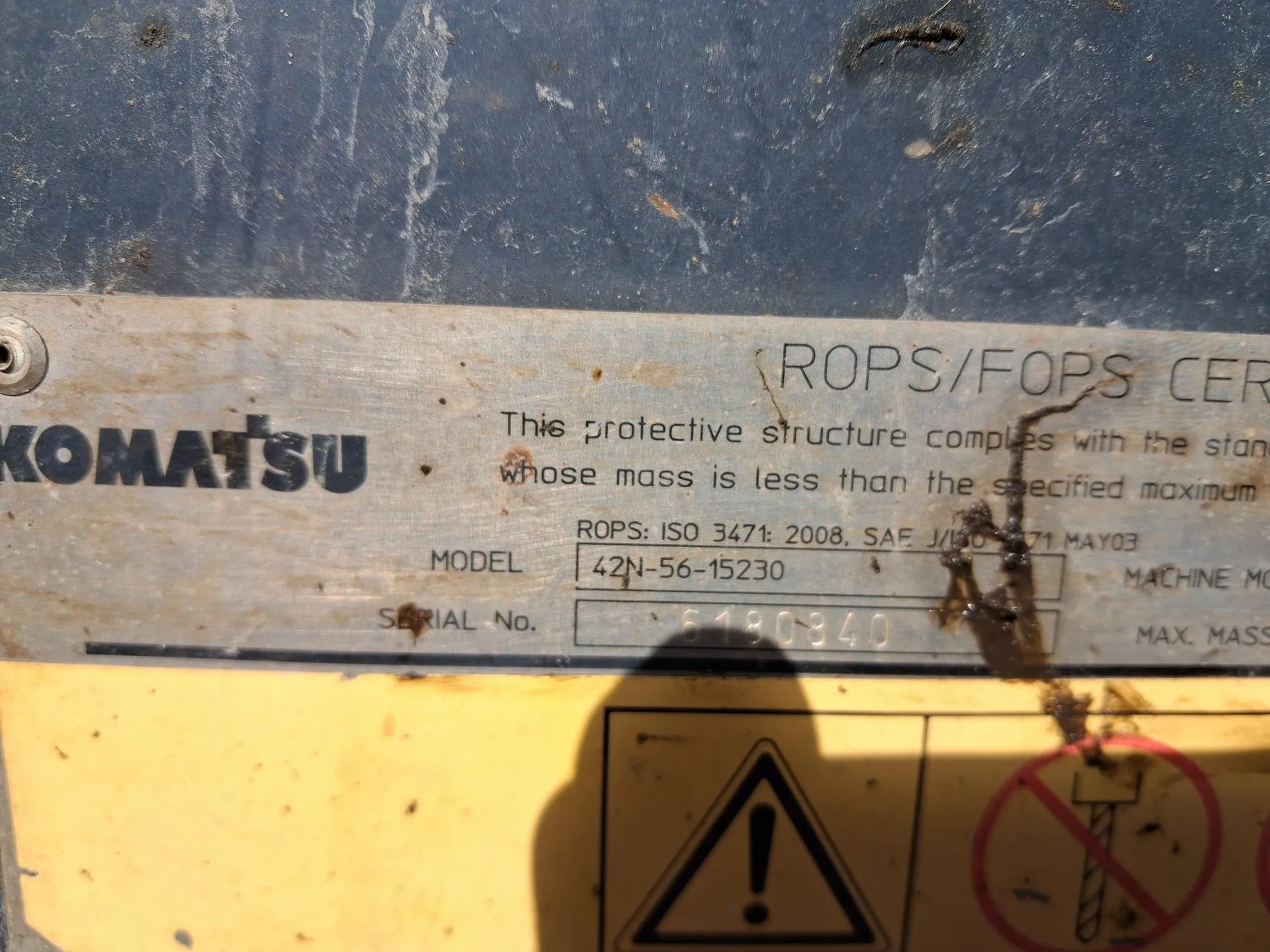Zdjęcie przedstawia metalową tabliczkę z napisem KOMATSU po lewej stronie. Na tabliczce widoczny jest tekst: ROPS/FOPS CER, This protective structure complies with the standard whose mass is less than the specified maximum, ROPS: ISO 3471: 2008, SAE J/ISO 3471 MAY03, MODEL 42N-56-15230, SERIAL No. 6180840. Poniżej tabliczki znajduje się żółta powierzchnia z dwoma znakami ostrzegawczymi: trójkątem z wykrzyknikiem oraz czerwonym kołem z przekreślonym symbolem wiertarki. Tło tabliczki jest niebieskie z widocznymi śladowymi uszkodzeniami.