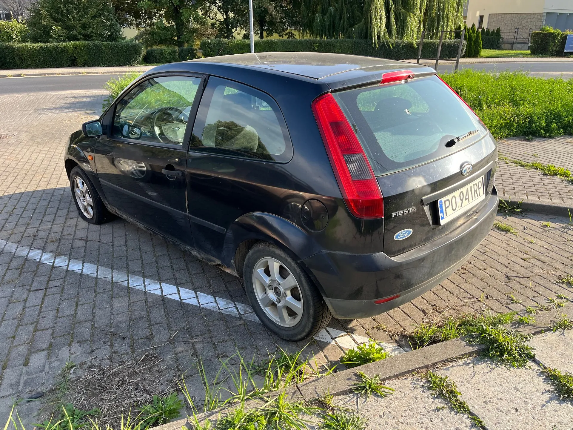 Czarny samochód osobowy marki Ford Fiesta zaparkowany na kostce brukowej. Pojazd jest widoczny od tyłu i z boku. Na tylnej klapie znajduje się napis 'FIESTA', logo Forda oraz polska tablica rejestracyjna o numerze PO 941RP. W tle widoczne są zielone krzewy, drzewa i fragment drogi.